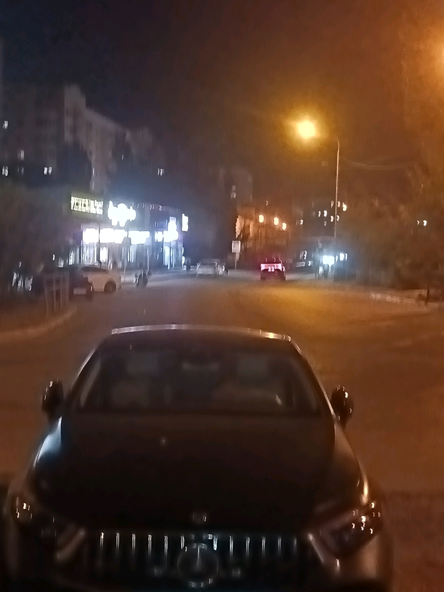 The image depicts a nighttime street scene in Astrakhan, Russia, likely a commercial or residential area given the presence of illuminated storefronts and apartment buildings. In the foreground, the hood and front grille of a dark-colored car are visible, suggesting the photo was taken from within another vehicle or at a very low angle. The street is illuminated by streetlights and the lights from buildings, creating a warm, hazy glow. Several other vehicles are present on the street, including a white car parked near a building with illuminated signs. In the mid-ground, a red car with its brake lights illuminated is visible, and a few indistinct figures are present near it. The overall impression is of a quiet, urban night.