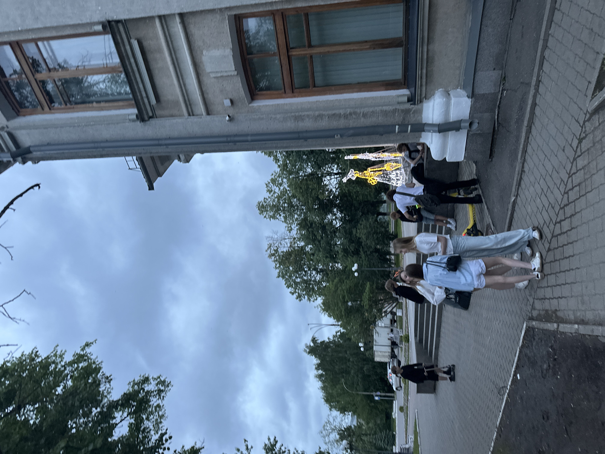 The image depicts a cloudy day in the city of Vologda, Russia, with a group of young people gathered in front of a building. The building appears to be made of concrete and features large, wooden-framed windows. To the left of the building, several trees and a street with a parked truck are visible. The ground is paved with interlocking bricks.
In the foreground, four young people are interacting. One person, wearing a light blue button-down shirt and white shorts, stands in the center, facing away from the camera. To their left, another person in a white shirt and light blue pants is also facing away. To the right of the central figure, a person in a white shirt is partially visible. Another person in a black shirt is seen on the left, standing near a small scooter.
In the background, a decorative Christmas tree illuminated with yellow lights stands prominently. More trees and a lamppost are also visible. The overall atmosphere suggests a casual gathering in an urban outdoor setting.