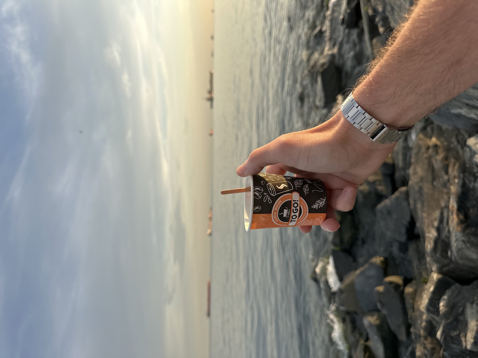 A person's hand, wearing a silver watch, holds a small paper coffee cup with an orange and black design that says "Coffee Break TO GO! PREMIUM QUALITY". The cup contains a wooden stir stick. The background shows a body of water with several large ships in the distance, under a cloudy sky with the sun setting, casting a warm glow. The foreground is composed of large rocks along a shoreline, with some small waves lapping at them. This scene is located in Bakırköy, Turkey, a district known for its coastline along the Sea of Marmara.