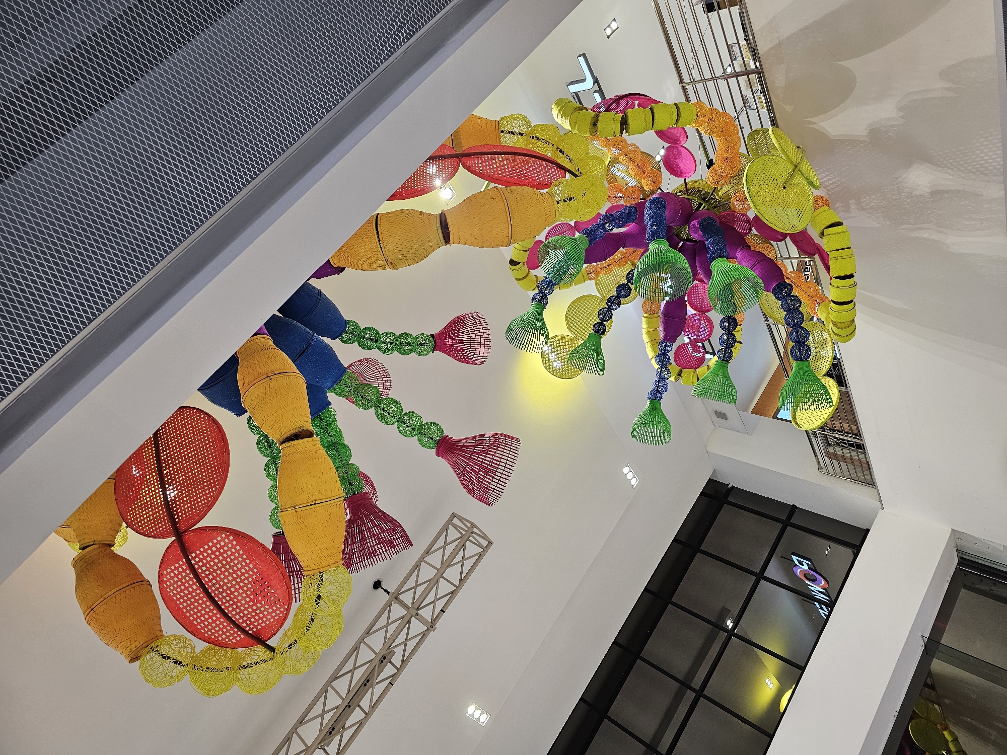 The image shows a large, colorful, abstract sculpture suspended from the ceiling of a modern building. The sculpture appears to be made of woven materials and features a variety of shapes and sizes, including spheres, cylinders, and flat discs. The dominant colors are yellow, red, blue, green, purple, and orange. The sculpture is hanging in an atrium or large open space, with a white ceiling and walls. In the background, a large window with dark framing reflects a lit sign that reads "BOM." A metal railing and stairs are visible on the right side of the image, suggesting an upper level or mezzanine. A section of a metal grate ceiling or ventilation system is visible in the upper right corner. The lighting in the space appears to be a mix of natural light from the window and artificial lighting from overhead fixtures, with a warm yellow light emanating from a light source in the upper center of the image. This installation is located at the Siam Center shopping mall in Bangkok, Thailand.