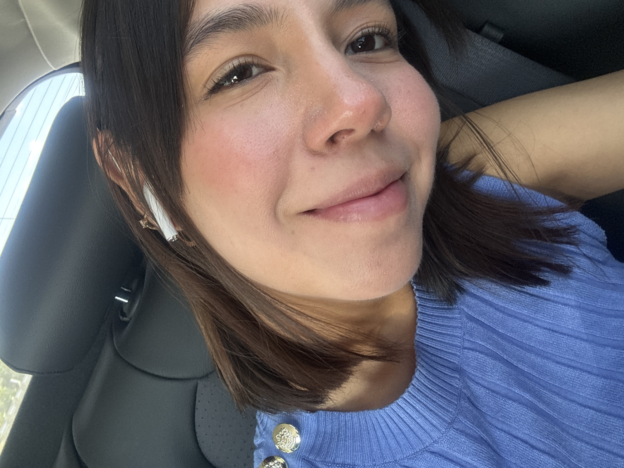 The image displays a close-up, angled self-portrait of a young individual inside a vehicle. The precise location within New Braunfels, United States of America, is not discernible from the visual evidence.
**Subject:** A young person with a light-to-medium complexion, dark brown, chin-length hair, and dark eyes. The individual is depicted with a slight smile and flushed cheeks, looking directly at the camera.
**Attire and Objects:** The person is wearing a light blue, ribbed knit top, with two golden, circular buttons featuring an embossed design visible on the left shoulder area. A white or light-colored earbud is present in the individual's right ear. A small, metallic hair clip is visible in the hair above the right ear.
**Environment:** The background consists of a dark gray or black car seat, including a headrest, indicating an interior vehicle setting. A car window in the upper portion of the image shows a bright, clear sky with faint green foliage, suggesting daytime conditions. The lighting is natural and bright.
**Action:** The individual is actively posing for a self-portrait within the vehicle.