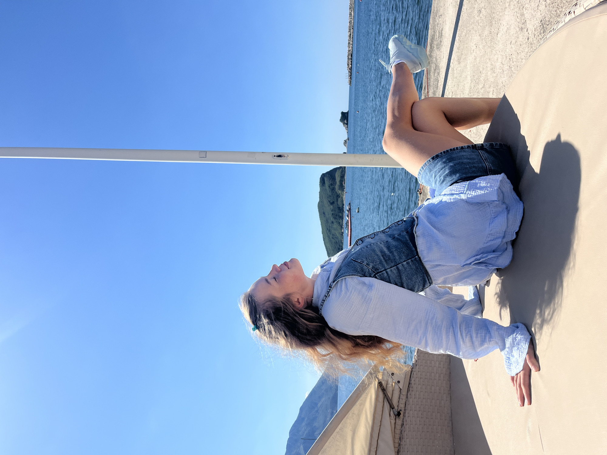 A girl rests in the sun at Budva, Montenegro. She is lying on her back atop a smooth, tan, curved surface; her head points toward the lower left of the frame, and her feet extend to the upper right. She wears denim shorts, a light blue button-up shirt, and a dark denim vest. The woman's hair is blonde and her skin is light-toned. Her legs are crossed at the ankles, and she wears white sneakers. The sky is a clear blue, with a thin, white pole running horizontally across the frame. A body of water with a distant island and boats stretches out behind her. A partial view of a canvas structure appears in the lower left corner. The scene appears to be outdoors in bright daylight.