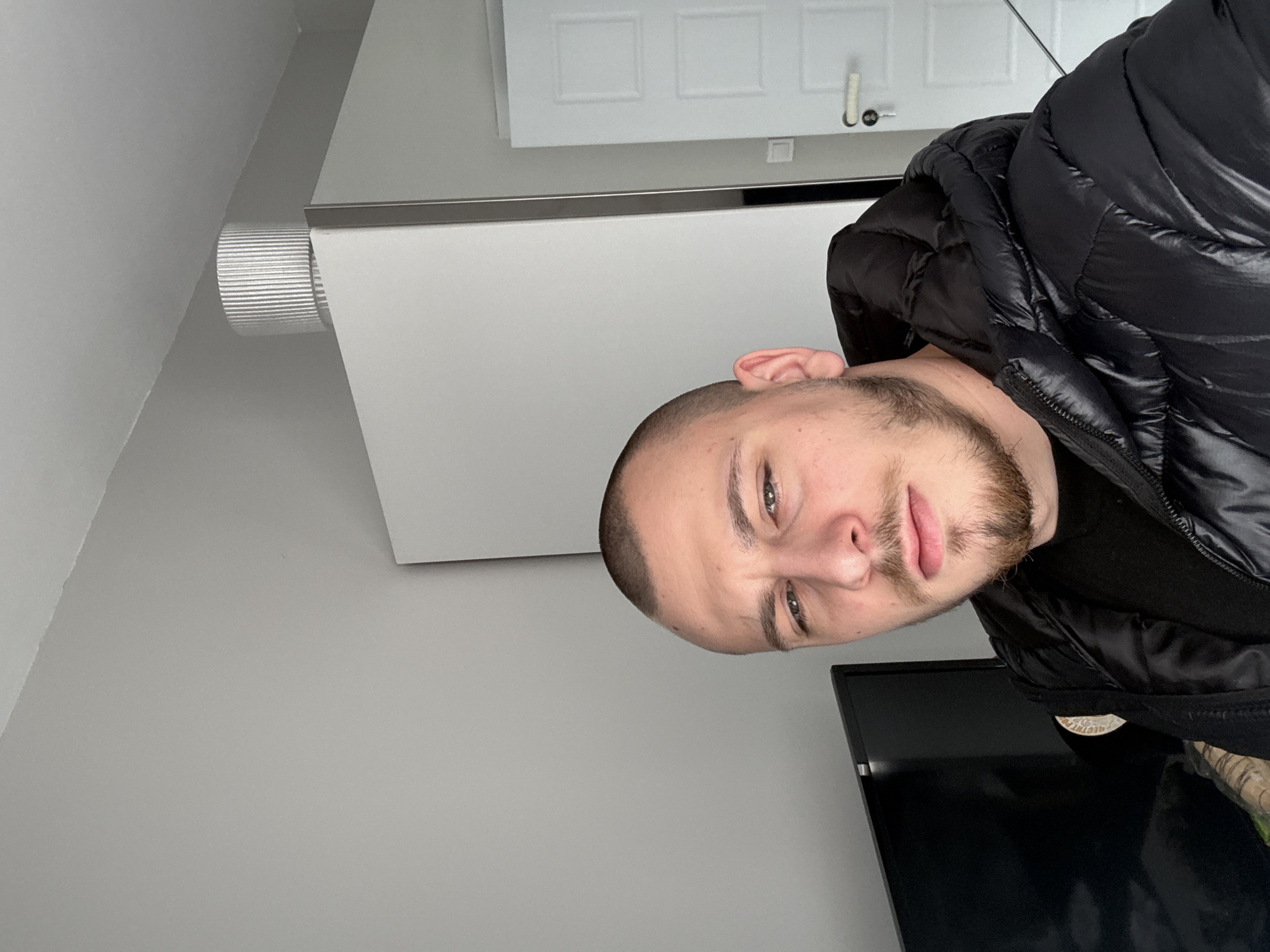 A portrait shot of a man with short brown hair and a trimmed beard, wearing a black puffy jacket. He is positioned at an angle, with a light gray wall visible behind him. A white radiator is above him, with a section of a white door in the top background. Part of a black television screen is visible in the bottom right. The location cannot be determined from the image.