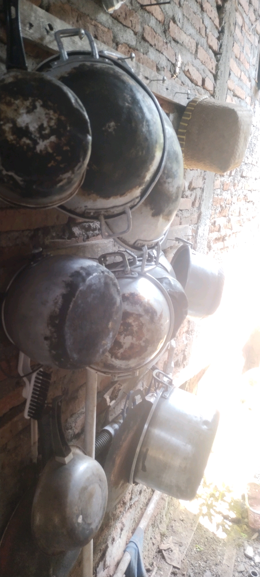 In a low-angle shot taken outdoors in Daleman, Indonesia, a collection of used and worn cooking pots and pans are seen hanging from a wooden plank attached to a brick wall. The pots vary in size and material, with some appearing to be made of aluminum or stainless steel, exhibiting signs of rust and discoloration from use. They are arranged in a tiered fashion, with larger pots suspended above smaller ones and some appearing to be stacked or nested. To the right, a woven basket with a distinctive pattern is also visible, possibly used for storage or as a lid. A white brush, possibly a cleaning tool, is attached to the side of one of the pots. The brick wall behind the pots shows some signs of age and wear. The lighting in the scene is harsh and bright, suggesting it may be daytime, with a strong light source coming from the right side of the frame, creating lens flare and washing out details in that area. The overall impression is one of a utilitarian outdoor kitchen space, likely in a rural or less developed setting, where cooking equipment is stored efficiently.