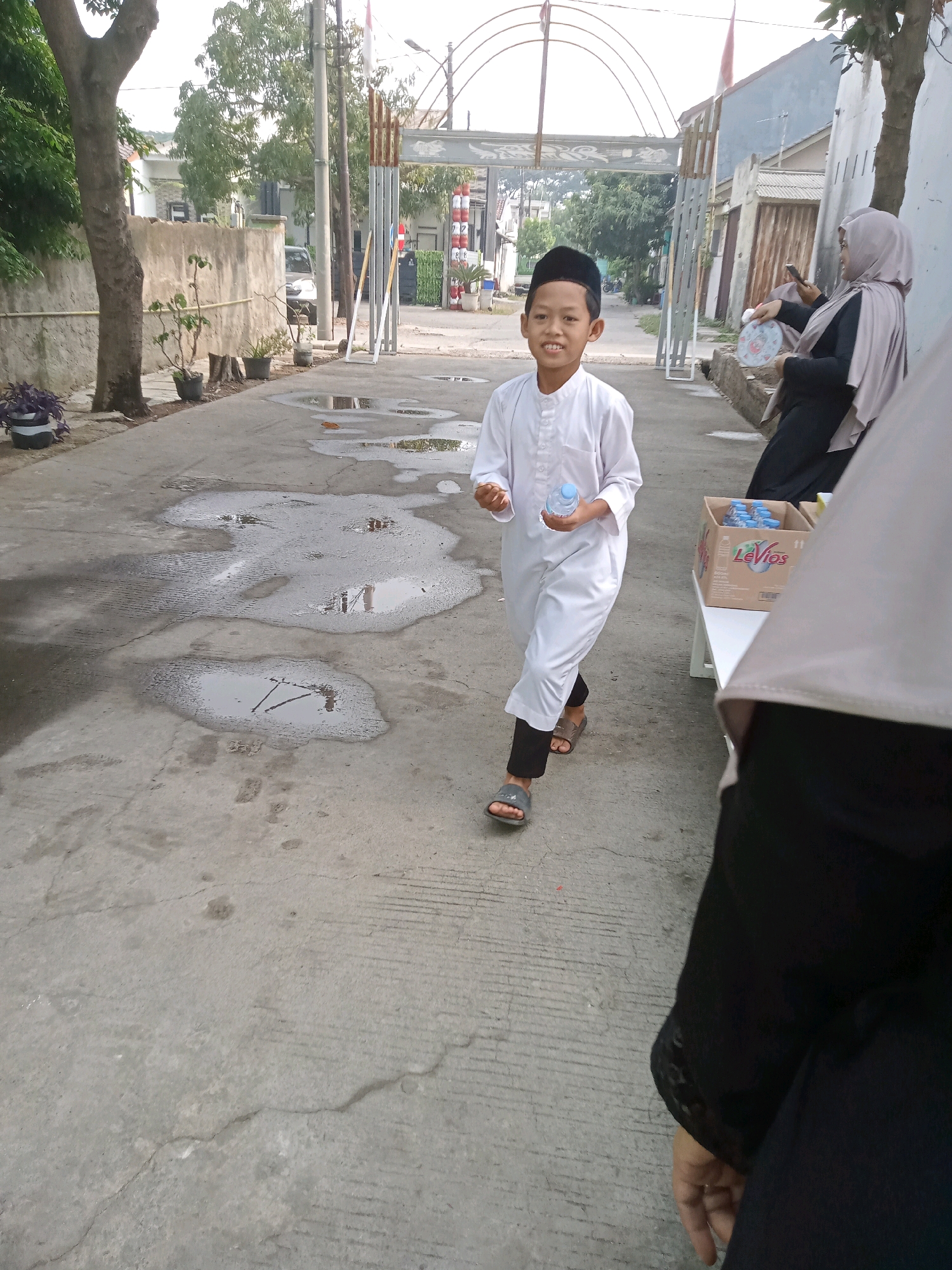 A young boy wearing a white peci and white traditional clothing is walking on a wet concrete pathway. He is holding a bottle of water in his right hand and looking at the camera with a smile. The boy is dressed in a Muslim outfit, consisting of a long-sleeved white tunic and dark pants. He is also wearing open-toed sandals. The pathway is lined with trees and plants on the left side, and buildings on the right. A woman in a hijab is sitting on a bench on the right side of the path, holding a fan and looking at her phone. A box of bottled water is on a table next to her. In the background, there is an archway and buildings. The sky is overcast. The scene appears to be in a residential area of Karawang, Indonesia.