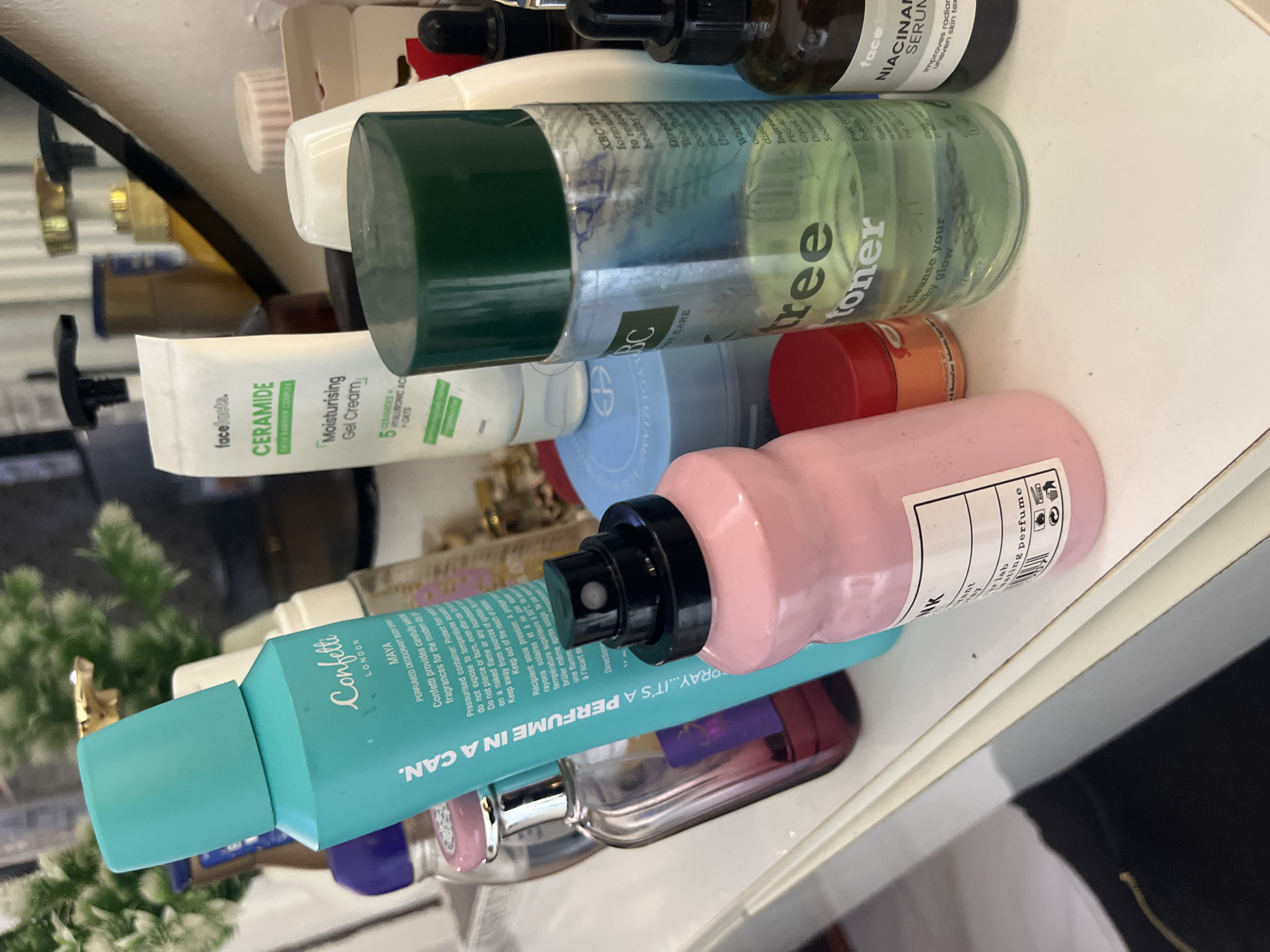 This is a vertical, close-up shot of a collection of skincare and beauty products, likely arranged on a shelf or dresser. The products are varied in size, shape, and color, creating a visually busy composition. Prominent items include a large green-capped toner bottle with "tree toner" visible, a pink spray bottle, a teal aerosol can labeled "Confetti" and "MAYA LONDON," a white tube of "facefacts. CERAMIDE Moisturising Gel Cream," and a dark glass bottle with a dropper, identifiable as "NIACINAM SERUM."
The lighting appears to be artificial and bright, casting some shadows. The background is out of focus but suggests an indoor setting, possibly a bathroom or vanity area. There are no people visible in the frame. The overall impression is one of a personal collection of beauty items, possibly undergoing organization or in active use. The text on the labels indicates various skincare benefits and ingredients like ceramides, hyaluronic acid, and tea tree.
The scene is set in Abuja, Nigeria. There are no discernible cues related to the time of day or weather. The arrangement of products suggests a domestic setting where personal grooming is a priority.