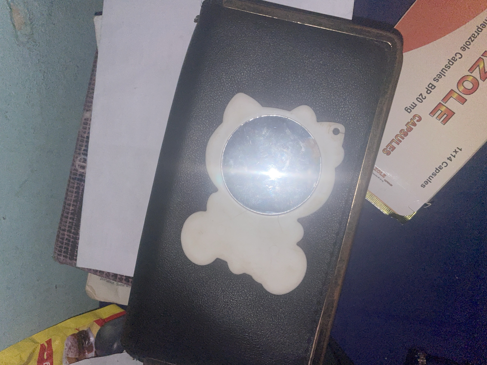 A white, cartoon-shaped keychain with a circular mirror in the center rests on a black, textured surface, possibly a wallet or pouch. The background includes a light blue, textured wall, stacks of paper, and a yellow and red packaging. Below the black surface is a box of Omeprazole Capsules BP 20 mg, with the text "1x14 Capsules" and "ZOLE CAPSULES" visible. The lighting is harsh, causing a bright reflection on the mirror. The scene appears to be indoors, on a flat surface, with an object and medical packaging. No people or animals are visible. The specific location in Jalingo, Nigeria, cannot be determined from the image.