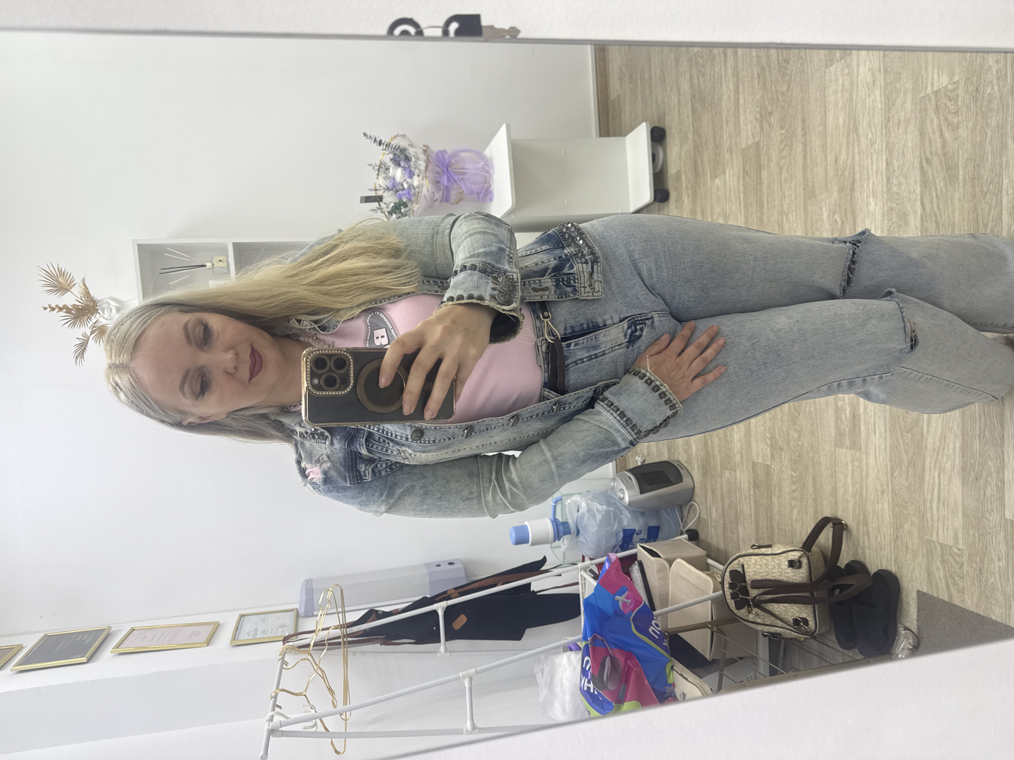 The image shows a woman taking a mirror selfie in what appears to be a beauty salon or retail space in Komsomolsk-on-Amur, Russia. The woman is wearing a light-wash denim jacket over a pink t-shirt and distressed denim jeans. She is holding a smartphone in her right hand, which is angled to capture her reflection. The background of the image reveals several elements of the environment. To the left, there is a clothing rack with hangers and some bags on the floor. Further back, a white shelf unit holds decorative items, including a vase with purple flowers and a diffuser. On the floor, near the woman, are a black backpack and a pair of black sandals. The overall impression is a casual, personal snapshot taken within a commercial or service-oriented setting.