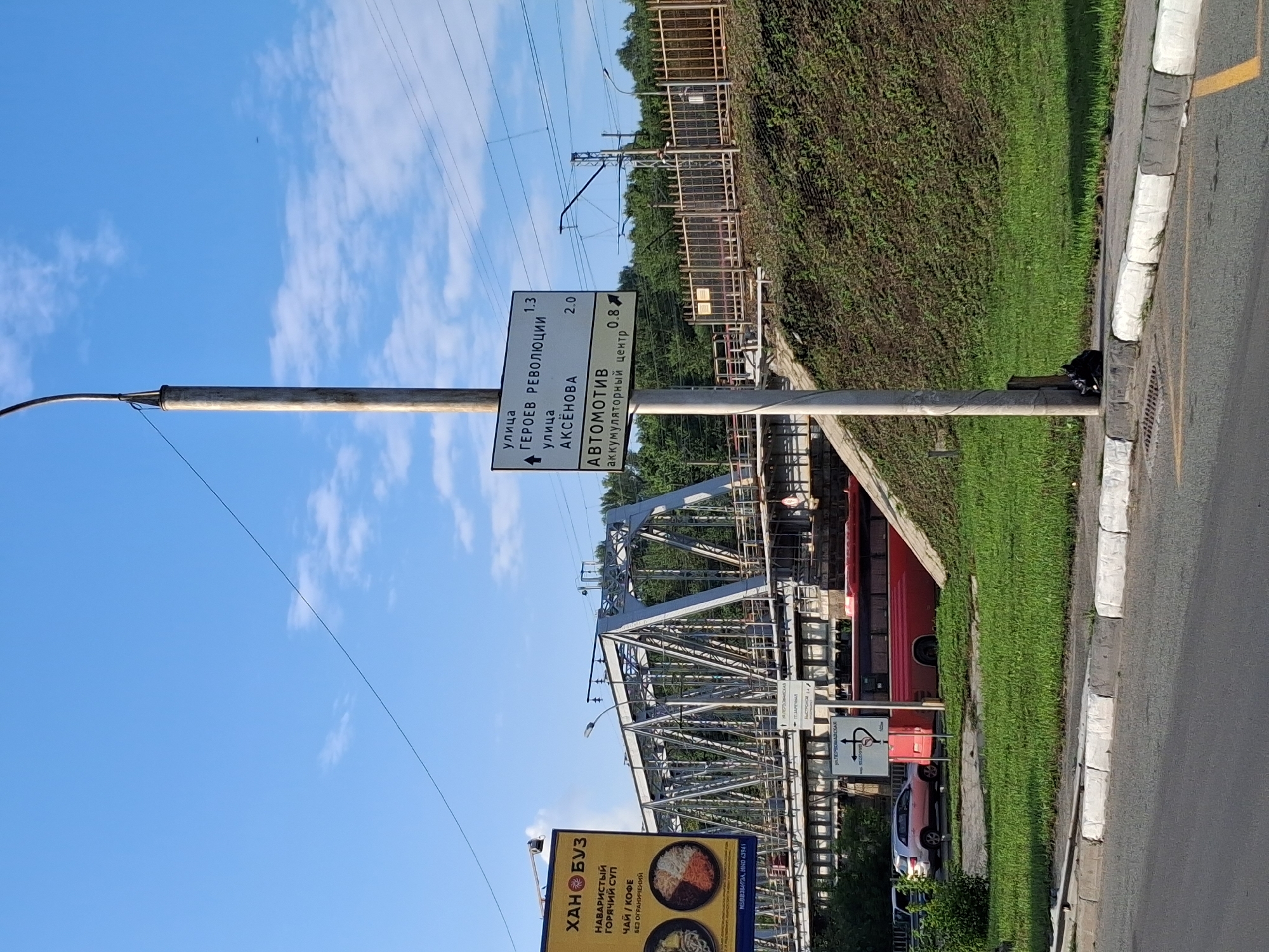The image captures an outdoor scene on a sunny day with a bright blue sky and some scattered clouds. A tall, weathered metal pole stands prominently in the foreground, supporting a white directional sign. The sign has text in Russian and indicates distances to various locations.
The sign reads:
"улица ГЕРОЕВ РЕВОЛЮЦИИ 1.3" (street Heroes of Revolution 1.3)
"улица АКСЁНОВА 2.0" (street Aksyonova 2.0)
"АВТОМОТИВ аккумуляторный центр 0.8" (Automotive battery center 0.8)
To the left of the pole, a section of a large metal railway bridge is visible. Underneath the bridge, a red bus is parked, and to its left, a light-colored SUV is parked on the side of the road. A billboard with yellow and black signage, featuring text in Russian and images of food, is also present on the left side of the frame.
The background consists of a grassy slope covered in dense green vegetation and trees. Beyond the slope, a metal fence separates it from what appears to be a railway line with overhead electrical wires and support structures. The ground in the immediate foreground is paved with asphalt and features a white-lined curb. A black trash bag is visible at the base of the metal pole. The overall impression is a view from a roadside, looking towards a railway bridge and urban infrastructure.