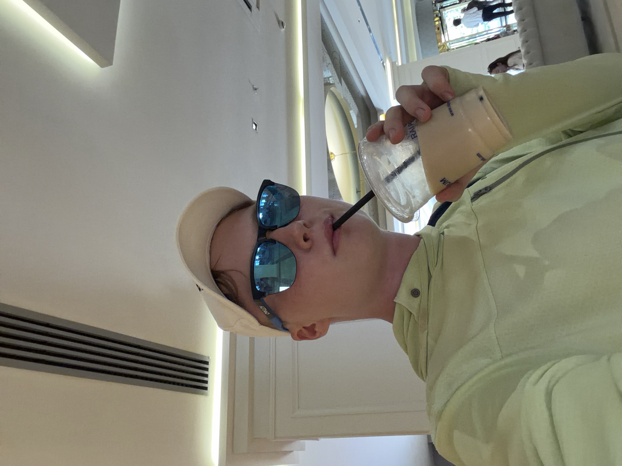 A young male wearing a light green hooded jacket, a white baseball cap, and blue mirrored sunglasses is holding a clear plastic cup with a straw and drinking from it. The cup appears to contain a light brown beverage. The background suggests an indoor setting with a modern ceiling featuring recessed lighting and air vents. The man is looking towards the camera. In the far background, there are hints of people and an entrance with natural light. The overall impression is of a casual moment captured in a public indoor space. Based on the provided context, this scene is located in the city of Kemer, Turkey.