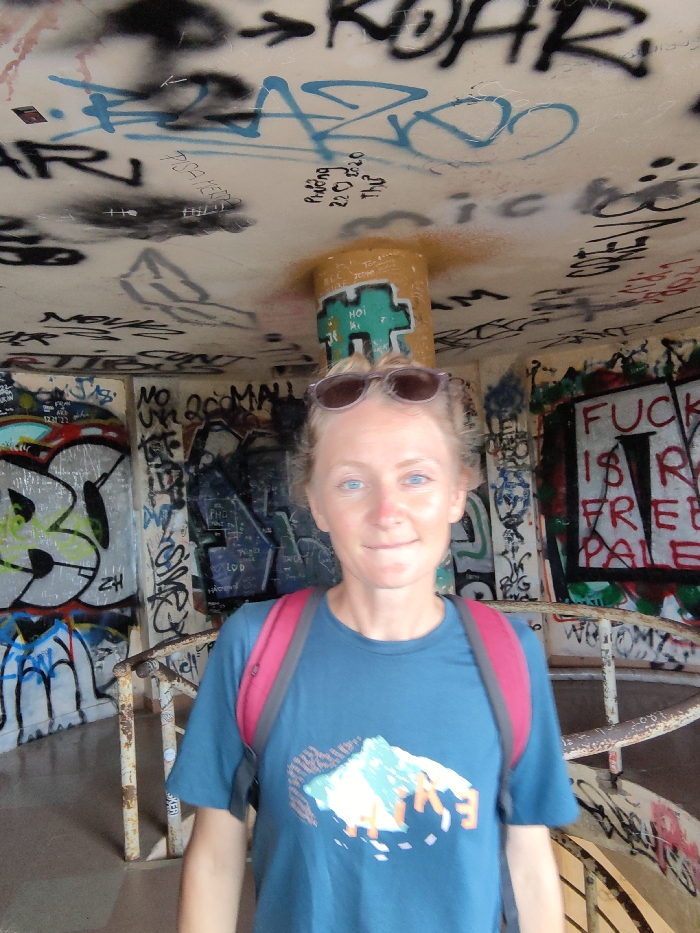 The image displays a woman positioned inside a heavily graffitied concrete structure, consistent with an abandoned military bunker or observation post, located in Thủy Xuân Ward, Vietnam. The subject, a fair-skinned woman with light hair and blue eyes, wears a blue t-shirt with a multi-colored mountain graphic and a red backpack, with sunglasses resting on her head. She looks directly at the camera with a faint smile. All visible surfaces—the rounded ceiling, concrete walls, and a central pillar—are entirely covered in layered spray-painted tags, drawings, and text. Prominent graffiti includes "ROAR", "PAZ", "Phuong 22.2.20", and "HOI AN" on the ceiling and pillar, and a large phrase "FUCK IS FREE PALE" on the right wall. A rusty metal spiral staircase with a matching handrail is visible descending on the right side of the frame.