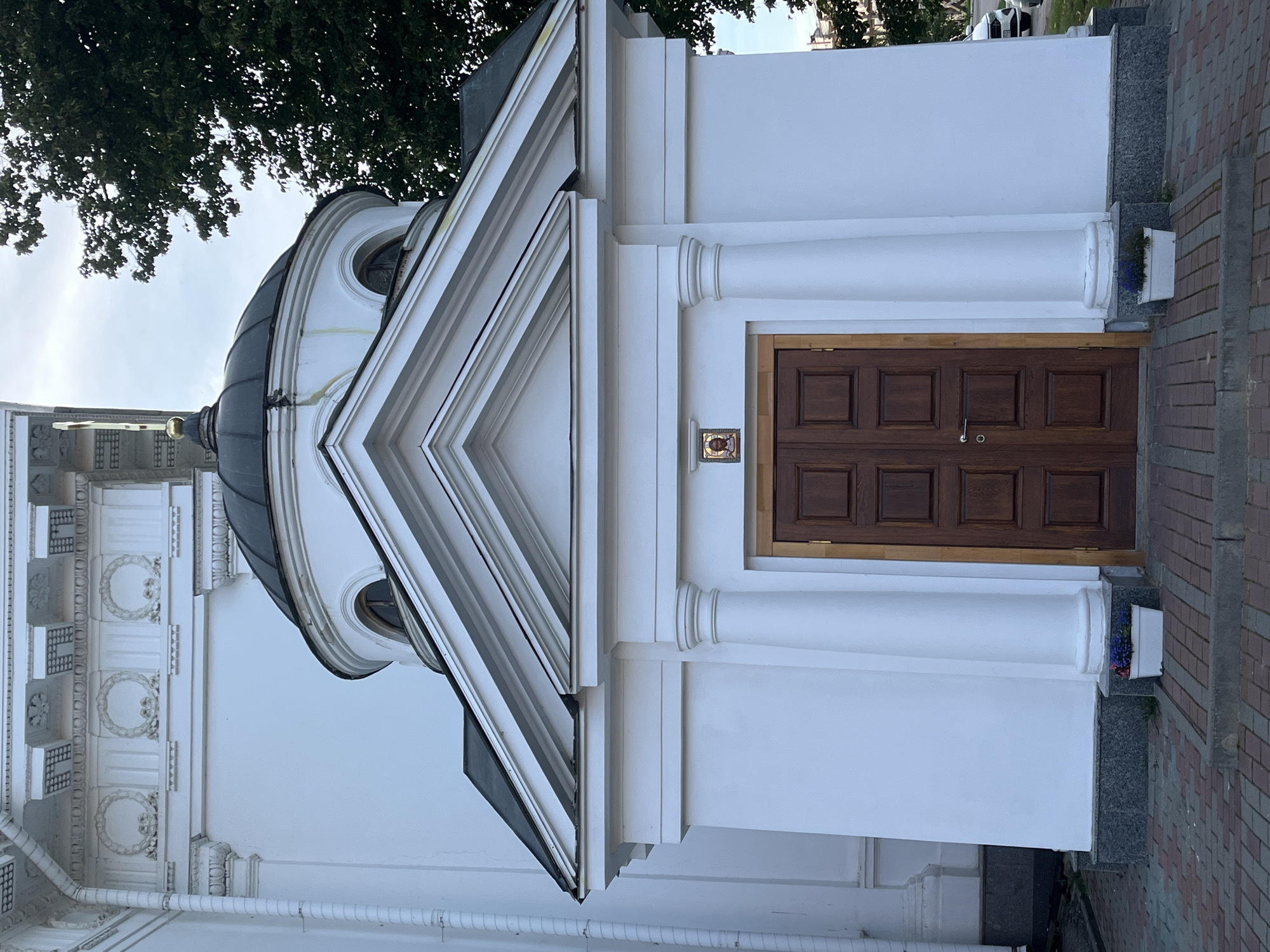The image shows a close-up, eye-level, exterior view of the entrance to a white building, possibly a church or chapel, in Nizhny Novgorod, Russia. The building features classical architectural elements such as fluted columns, a pedimented entrance, and a domed roof, which is dark in color. A detailed cornice with decorative moldings and circular motifs is visible on the upper part of the main building, to the left of the entrance. The entrance itself is a set of double wooden doors with a dark stain and square panels. Above the doors, a small religious icon is mounted. Two white planters with dark blue flowers are placed on either side of the entranceway, resting on a grey stone base. The ground in front of the entrance is paved with red and grey bricks. A large green tree is partially visible on the right side of the image, and the sky is overcast. A small portion of a white car is visible in the background on the right, suggesting the presence of a street or parking area. The overall impression is one of architectural detail and solemnity.