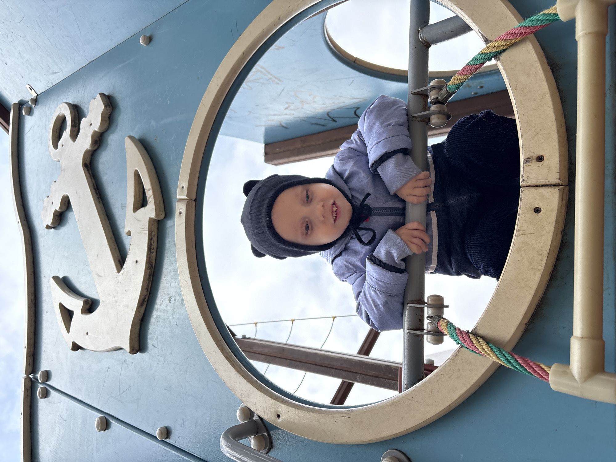 A low-angle, slightly rotated medium shot depicts a young child playing on a playground structure in the city of Chaikovsky, Russia. The child, wearing a blue jacket and a grey hat with small ears, is partially visible through a circular porthole. The child is grasping a vertical metal pole within the structure, appearing to look down at the camera with a slight smile. The playground structure is painted in shades of blue and cream, and features a decorative anchor motif attached to the side. The background shows a glimpse of the sky and other parts of the playground equipment. The photo is taken outdoors.