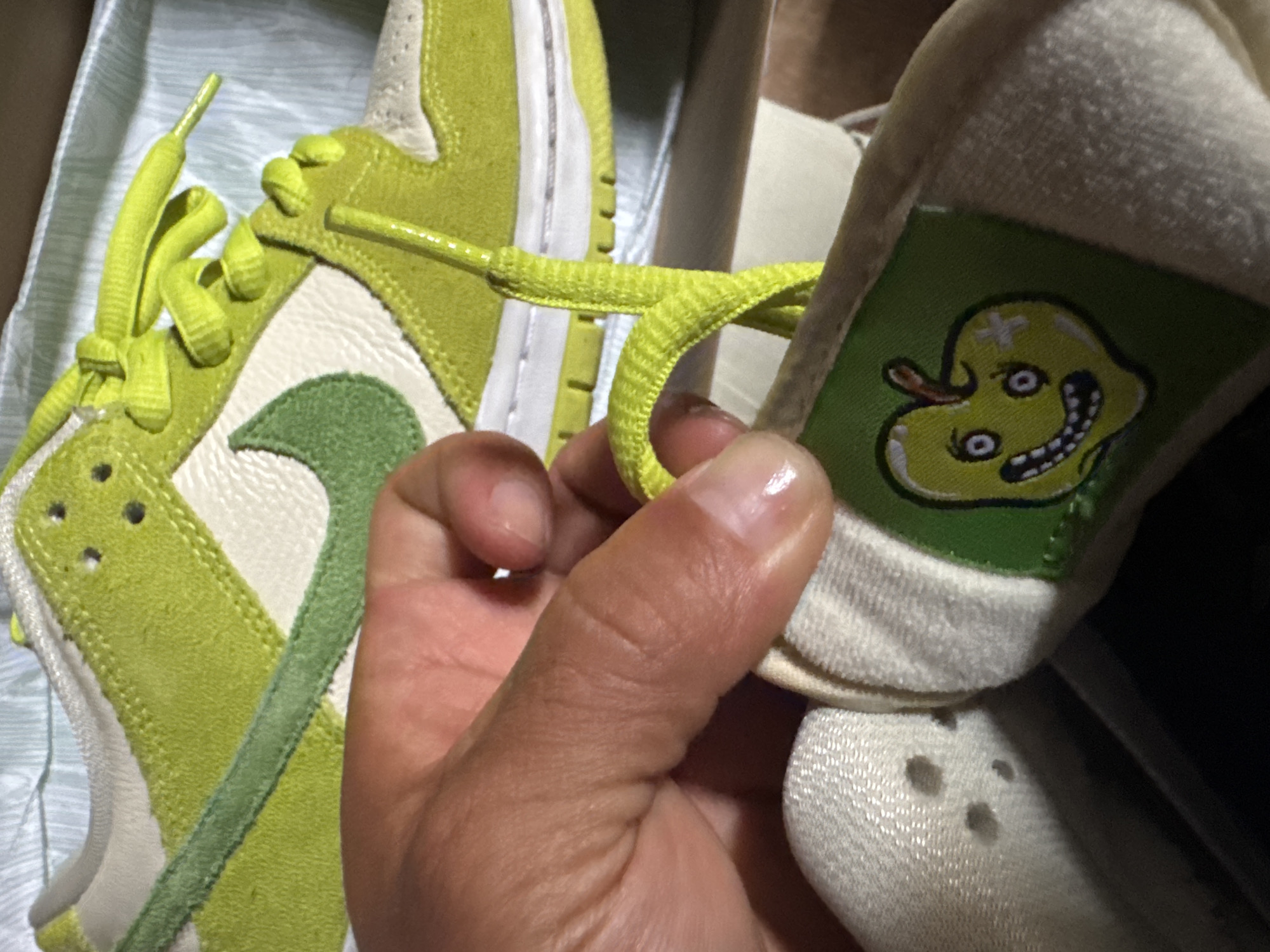 A hand is holding up a sneaker. The sneaker is a low-top model, primarily light green and off-white. The side has a light green swoosh symbol. The tongue has a green patch with an illustration of an anthropomorphic yellow apple with a sinister face. Another nearly identical shoe is partially visible in the background, oriented towards the left of the frame. Both shoes appear to be new, possibly still in a box. The environment suggests an indoor setting.