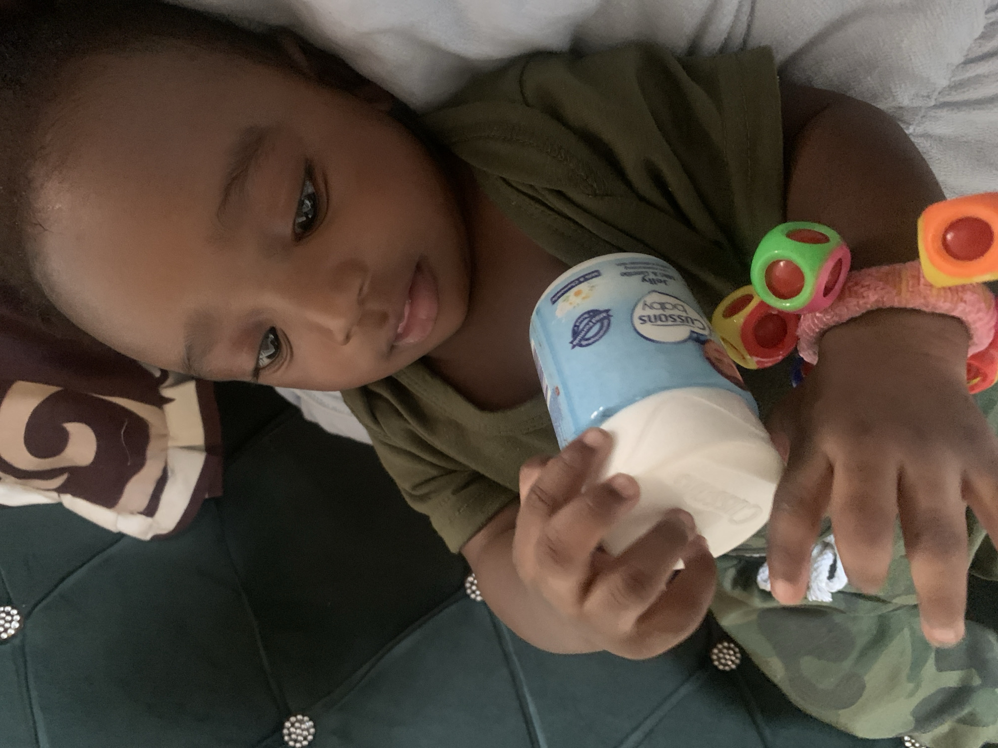 A young child lies on what appears to be a dark green tufted surface, possibly a couch or bed, covered with bedding. The child is wearing a dark green shirt and camo pants and holding a blue and white baby product container. The child also wears a colorful toy on their wrist.
There is no information in the image that indicates this scene's location in Barakin Akawo, Nigeria. Therefore, it's impossible to confirm the location.