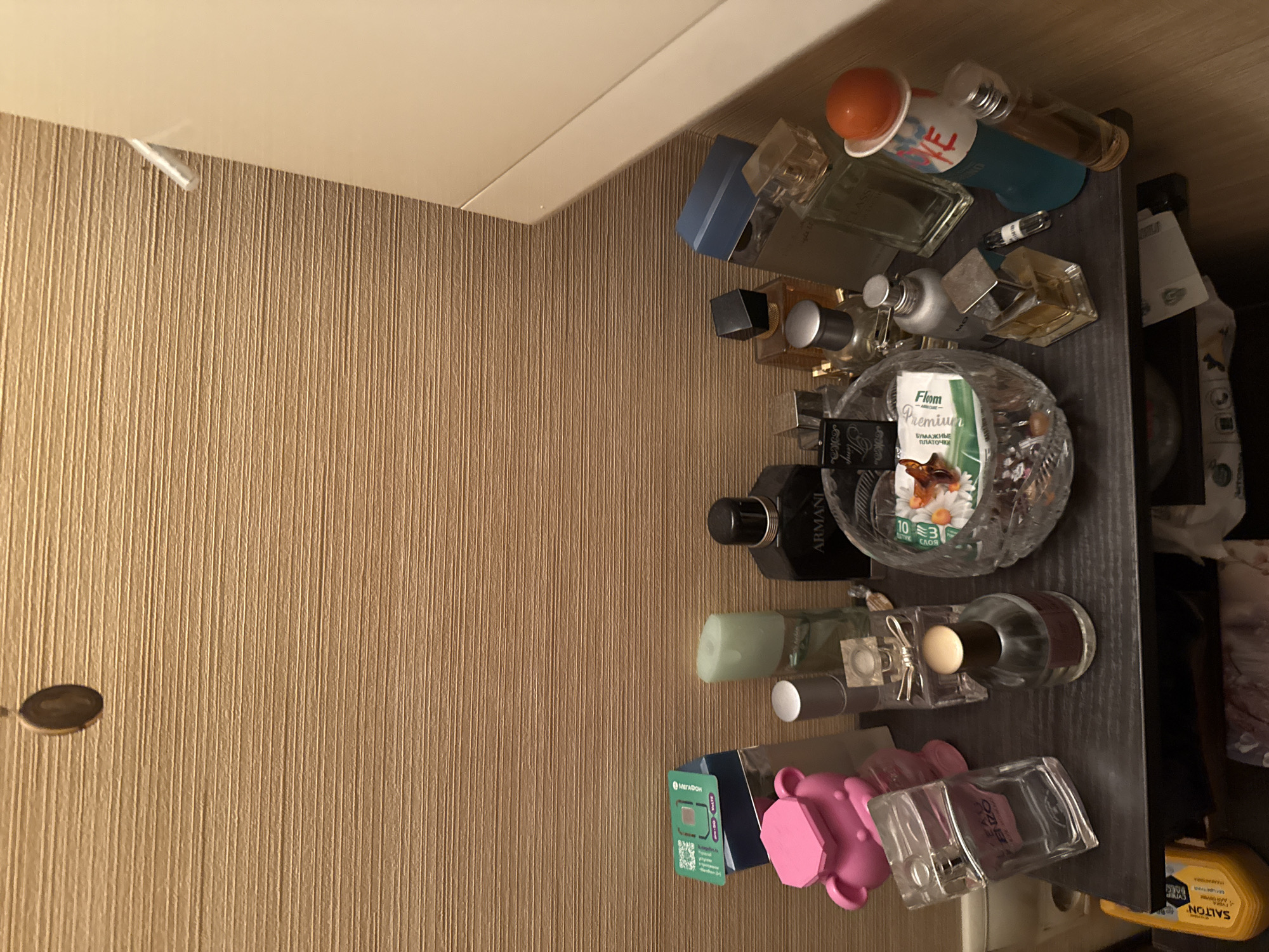 This is a vertical, eye-level shot of a collection of perfumes on a shelf. The shelf is dark, with the items arranged on it. The background is a textured, light brown wall.
The objects on the shelf include several perfume bottles of various shapes, sizes, and colors. Some bottles are clear glass, while others are opaque. There is a distinctive crystal perfume bottle with flowers depicted on it, and a small green box with Russian text that appears to be "MEGAPHONE." There are also a couple of pink bear-shaped objects.
The lighting suggests it may be indoors, possibly in a bathroom or dressing area. The time of day and weather are not discernible from the image.
Visible text includes "ARMANI," "Flom Premium," "БУМАЖНЫЕ ПЛАТОЧКИ," "MEGAPHONE," "SALTON," and "ENZO." There is also some text on a yellow container that appears to be "SHAMPAGNORA."