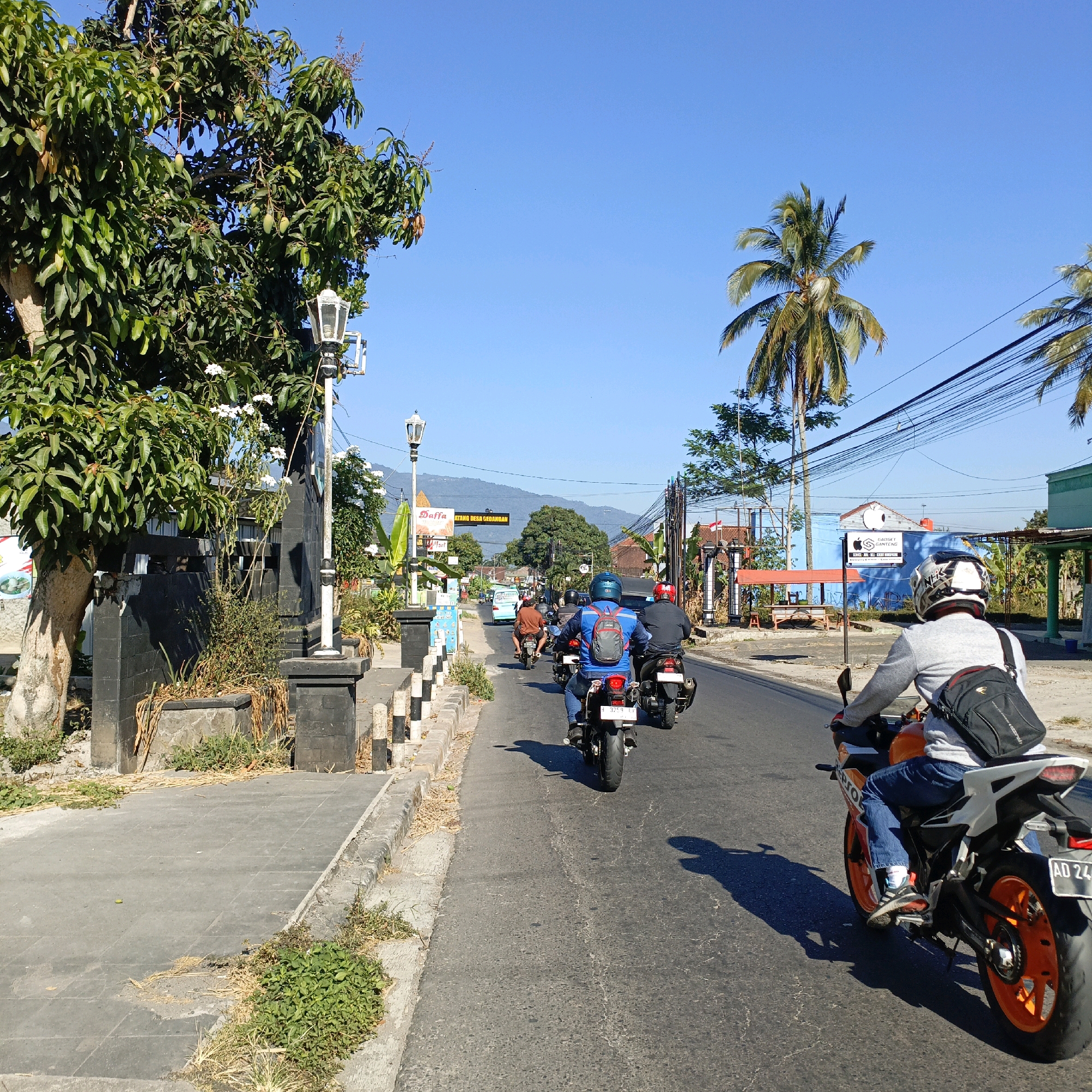 The image depicts a street scene in Gedangan, Indonesia, under a clear blue sky. The perspective is from a motorcycle, looking forward along a paved road. Several other motorcycles are visible ahead, also traveling in the same direction. The rider in the foreground is wearing a grey long-sleeved shirt, blue jeans, and a helmet with black and white markings. A black backpack is worn over the shirt. The motorcycle is orange and white with black accents. In the background, there are buildings, trees including a prominent palm tree, power lines, and hills or mountains in the distance. A sign with yellow text, partially obscured, is visible on the side of the road, and it appears to say "DATANG DESA GEDANGAN." There is a sidewalk on the left side of the road with some vegetation and decorative pillars.