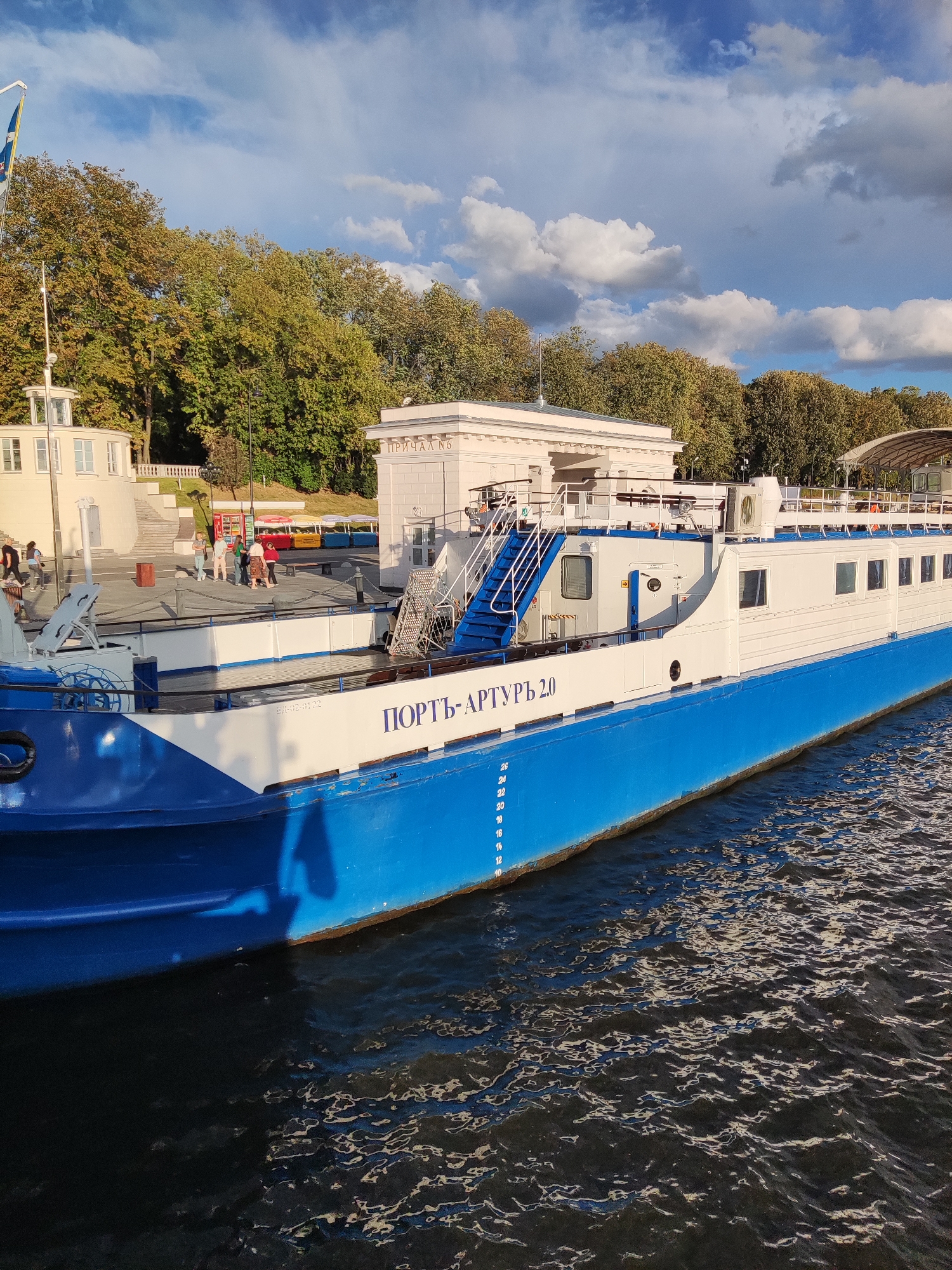 The image shows a blue and white boat docked at a pier on a sunny day. The boat has the Cyrillic text "ПОРТЬ-АРТУРЪ 2.0" written on its side. In the background, there is a white building with columns and a paved area with people walking. There are also trees with autumn foliage and a partly cloudy blue sky. The boat appears to be a passenger vessel. This scene is likely the Khimki River Port, located in Khimki, Russia.