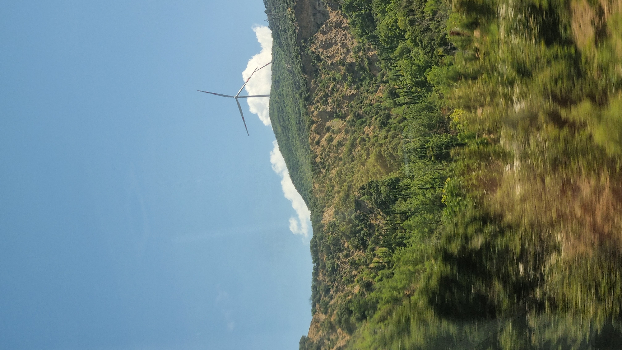 A lone wind turbine stands atop a green, tree-covered mountain under a bright blue sky with scattered clouds. The foreground is blurred, suggesting movement, and is filled with trees. The mountain itself has rocky, barren sections intermixed with lush vegetation.