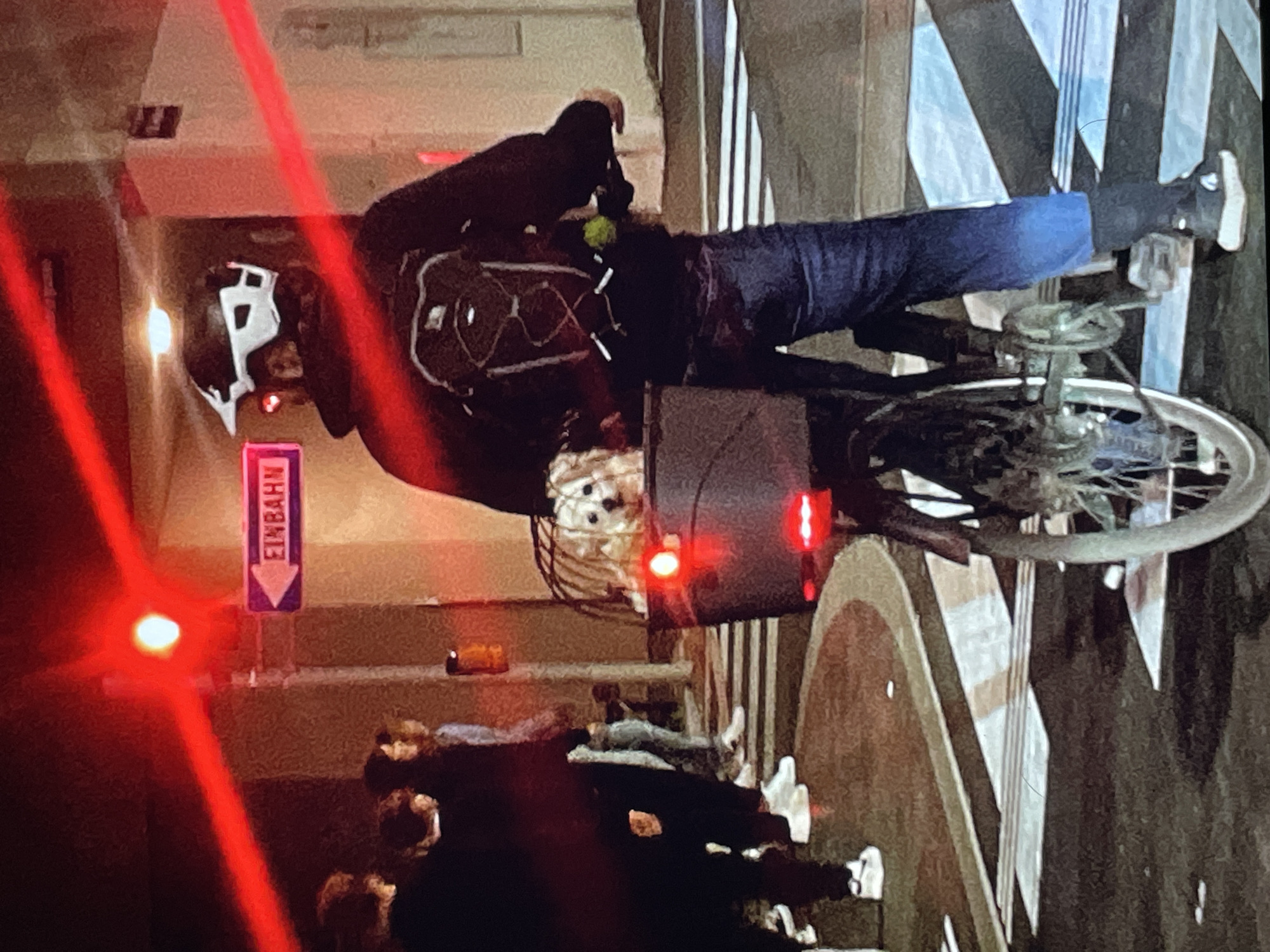 A cyclist wearing a helmet, dark clothing, and jeans is riding a bicycle. They have a backpack and a box attached to the back of the bike. The bicycle has a rear light. In the background, there is a group of people. A red laser beam shines across the scene, and a sign with the word "EINBAHN" (meaning "one way") is visible. The scene appears to be a street in a city.