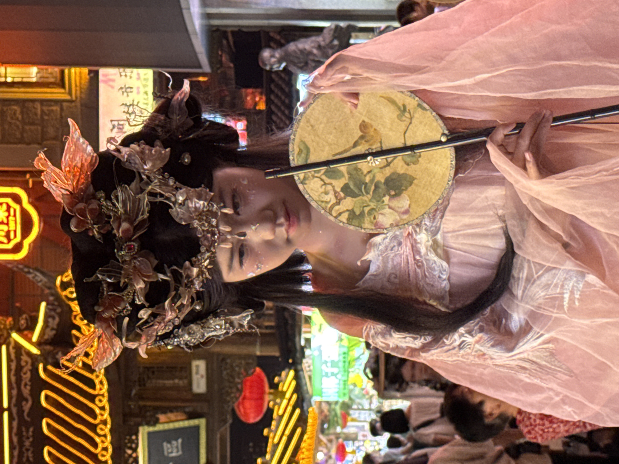 The image depicts a young woman dressed in traditional Chinese attire, likely a hanfu, adorned with an elaborate headdress featuring floral and leaf motifs and delicate beadwork. She has a light dusting of glitter on her cheeks. In her hands, she holds a round, decorative fan painted with a bird and floral design. She is partially obscured by a sheer pink overlay garment.
The background suggests a vibrant urban environment, possibly a historic street or market, in Yuzhong District, China. The scene is illuminated by bright, colorful neon lights and traditional red lanterns, indicating nighttime or a well-lit indoor setting. In the blurred background, other people can be seen, suggesting a public space. A statue of a person is visible in the far right background.
