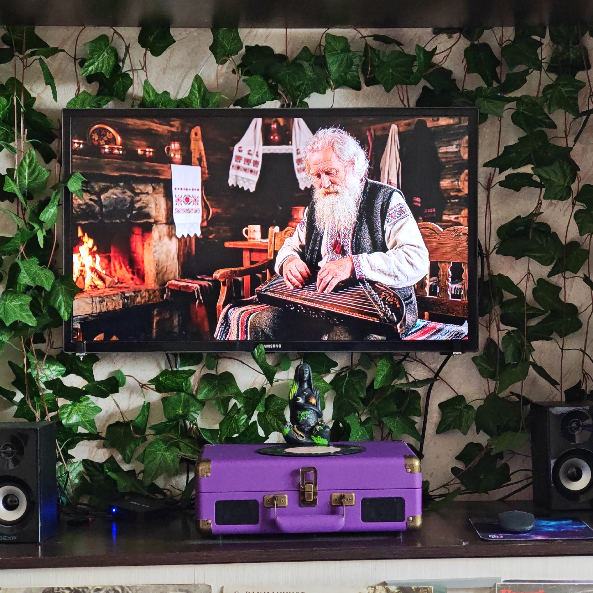 A television displaying a scene of an elderly man with a long white beard playing a stringed instrument in front of a fireplace is mounted on a wall adorned with artificial ivy. Below the television sits a purple suitcase-style record player, flanked by two black speakers. A statue stands on top of the record player, and a computer mouse rests on a mousepad to the right. The bottom of the frame reveals the edges of several record sleeves. The location appears to be a home interior and the scene is unlikely to be in Rostov, Russia because of the lack of information that it is in that city.