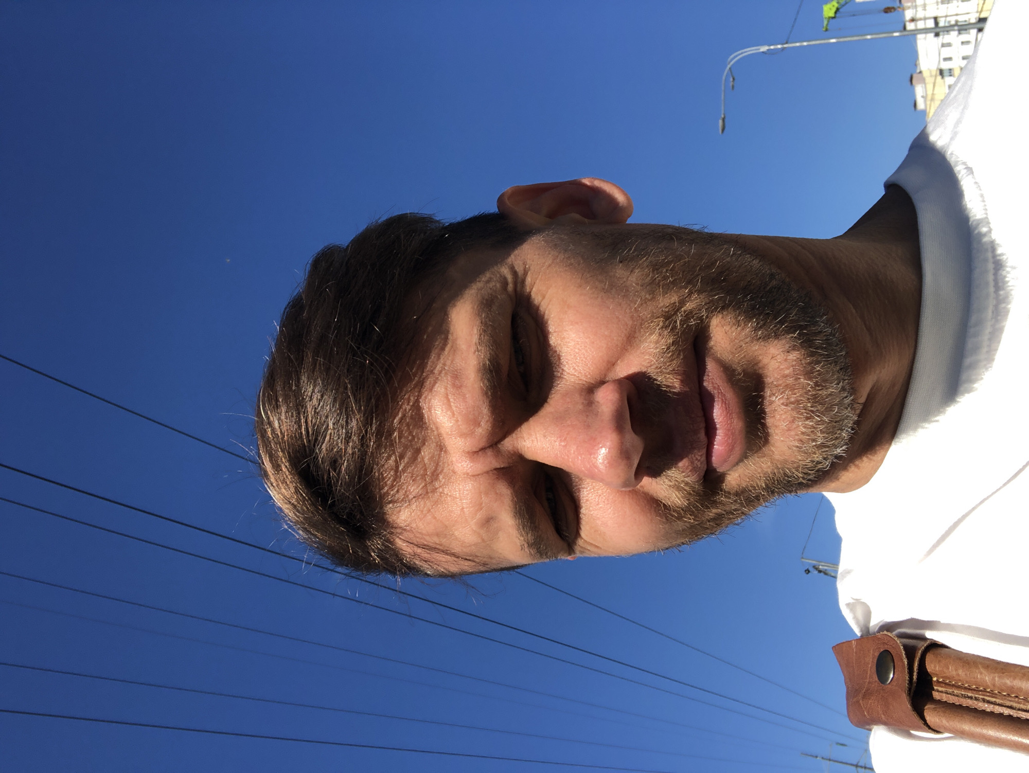 A close-up, low-angle selfie captures the face of a white man with short, light brown hair and a short, light brown beard. He is wearing a white t-shirt and a brown leather strap with a metal button is visible on his left shoulder. The background is a clear blue sky with several dark, parallel wires stretched across it. The man's face is lit by bright sunlight, casting shadows and highlighting the texture of his skin, including wrinkles on his forehead. To the right of his face, a portion of a streetlamp and a crane are visible against the sky, suggesting an urban environment. Based on the presence of parallel wires and the architecture of buildings that appear in the background, the location is likely St. Petersburg, Russia, possibly in a district with overhead power lines and ongoing construction.