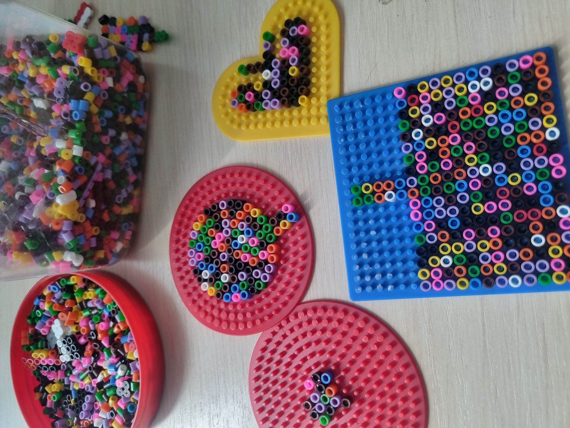 The image displays a collection of colorful fuse beads and pegboards, commonly used for crafting. In the foreground, there are three pegboards with beads arranged in various patterns. One is a circular red pegboard with a partial design of beads. Another is a larger circular red pegboard with a more complete circular pattern of beads. A rectangular blue pegboard is also visible, partially filled with beads forming an abstract design. To the right, a yellow heart-shaped pegboard has a design of beads. In the background, a red bowl is overflowing with loose fuse beads of various colors, and a clear plastic container also holds a large quantity of mixed beads. A small, completed fuse bead creation, possibly a figure, is seen to the right of the clear container. The objects are arranged on a light-colored, wooden surface. There are no people visible in the image. The location is not specified in the image.