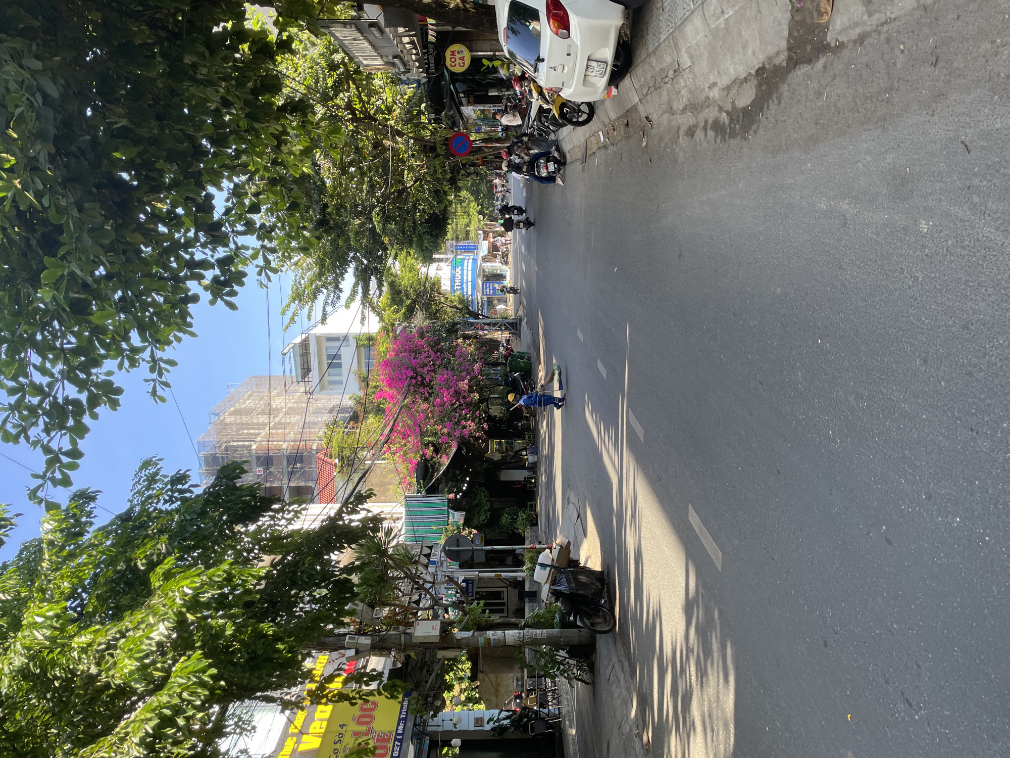 This is a street scene in Da Nang, Vietnam, on a sunny day with dappled light filtering through the trees. The street is paved with asphalt and has white lane markings. On the left side of the street, there are buildings with signs in Vietnamese, some with awnings and tables outside, suggesting cafes or shops. A person in a blue uniform and a yellow hard hat is sweeping the street with a broom. A parked motorcycle with a large trash bag tied to it is also on the left side. Further down the street, more buildings and parked motorcycles are visible.
On the right side of the street, there are more buildings with awnings and signs, including a "Barber Shop" and "COM GA." Several motorcycles are parked along the curb, and a white car is also parked there. A person is sitting on a motorcycle. The scene suggests a busy urban environment with a mix of commercial activity and residential buildings. The trees lining the street provide shade and add to the vibrant atmosphere of the city. The sky is clear and blue.