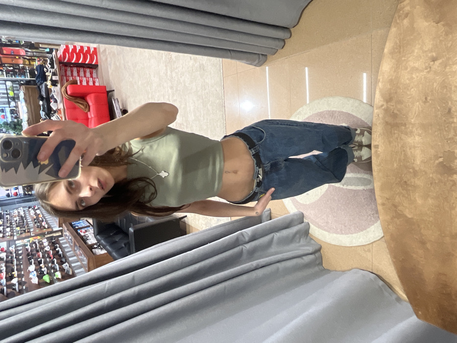 A young adult female is captured in a full-length mirror selfie within a retail establishment, specifically identified as a "Street Beat" sneaker and apparel store in Krasnoyarsk, Russia. The precise address within Krasnoyarsk is not discernible, but the store's interior design aligns with chain locations typically found in shopping malls.
The female, with long, straight dark brown hair, stands centrally in the image. She is dressed in an olive green sleeveless crop top, blue denim jeans with a black belt, and light-colored sneakers with dark laces. Her midriff is exposed, revealing a navel piercing. She holds a smartphone, partially obscuring her face, with her left hand, which is encased in a gray and blue mountain-patterned phone case. Her right hand is positioned at her right hip, with her thumb hooked into her jeans.
The background, a reflection in the mirror, shows the store's interior. To the left, a wall displays numerous pairs of sneakers on tiered shelves. Beneath these shelves is a dark wooden counter. In the mid-ground, a black two-seater sofa is visible, and further right, a bright red armchair is present, surrounded by stacks of red and white shoe boxes. Additional merchandise displays line the deeper background. The floor is covered with light-colored tiles, featuring a large circular rug with a distinctive light pink/beige and white stylized pattern. Gray fabric curtains, suggestive of a fitting room, frame both sides of the mirrored scene. The bottom edge of the image shows a wooden surface in the immediate foreground.