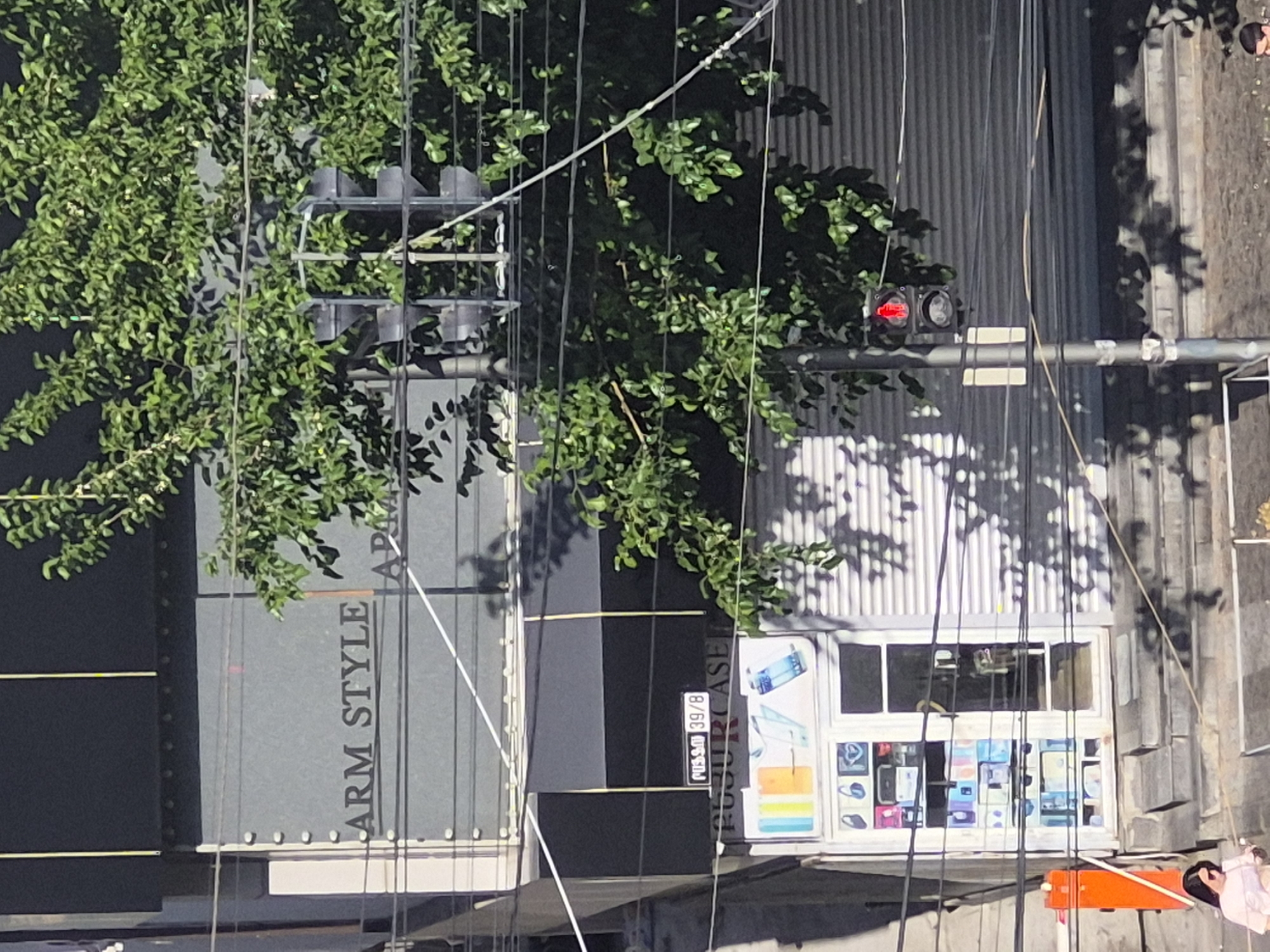 This elevated view captures a sunny urban street scene in Yerevan, Armenia, characterized by buildings, lush green trees, and a network of overhead wires. The image is oriented vertically, but when viewed upright, reveals a busy commercial area.
To the left, a tall building or wall features the partially visible text "ARM STYLE AP..." arranged vertically. A dense green tree stands prominently, its foliage partially obscuring the building and casting dappled shadows.
Emerging from the tree's branches is a traffic light, illuminated red, signifying a stop. Numerous black and white overhead cables crisscross the frame, indicative of a city environment.
Further right, a storefront is visible, its window showcasing a colorful display of electronic items, likely mobile phones or accessories like cases. Above the shop window, a small black sign displays the text "NISSAN 39/8," with additional, fainter text underneath that appears to read "FUGU CASE" or "FUGU GLASS," suggesting the nature of the business. The building facade behind the shop window is made of corrugated metal, reflecting bright sunlight and showing clear shadows from the overhead wires and tree branches.
In the bottom right corner, a person is partially visible, clad in a light-colored top, seemingly interacting with an orange object. Another person's head, possibly wearing a hat, is also partially discernible further to the right on the pavement.
The scene is bathed in bright daylight, evidenced by the strong, distinct shadows cast on the buildings and ground, suggesting a clear, sunny day, likely around mid-day. The overall impression is that of a typical, active city street.