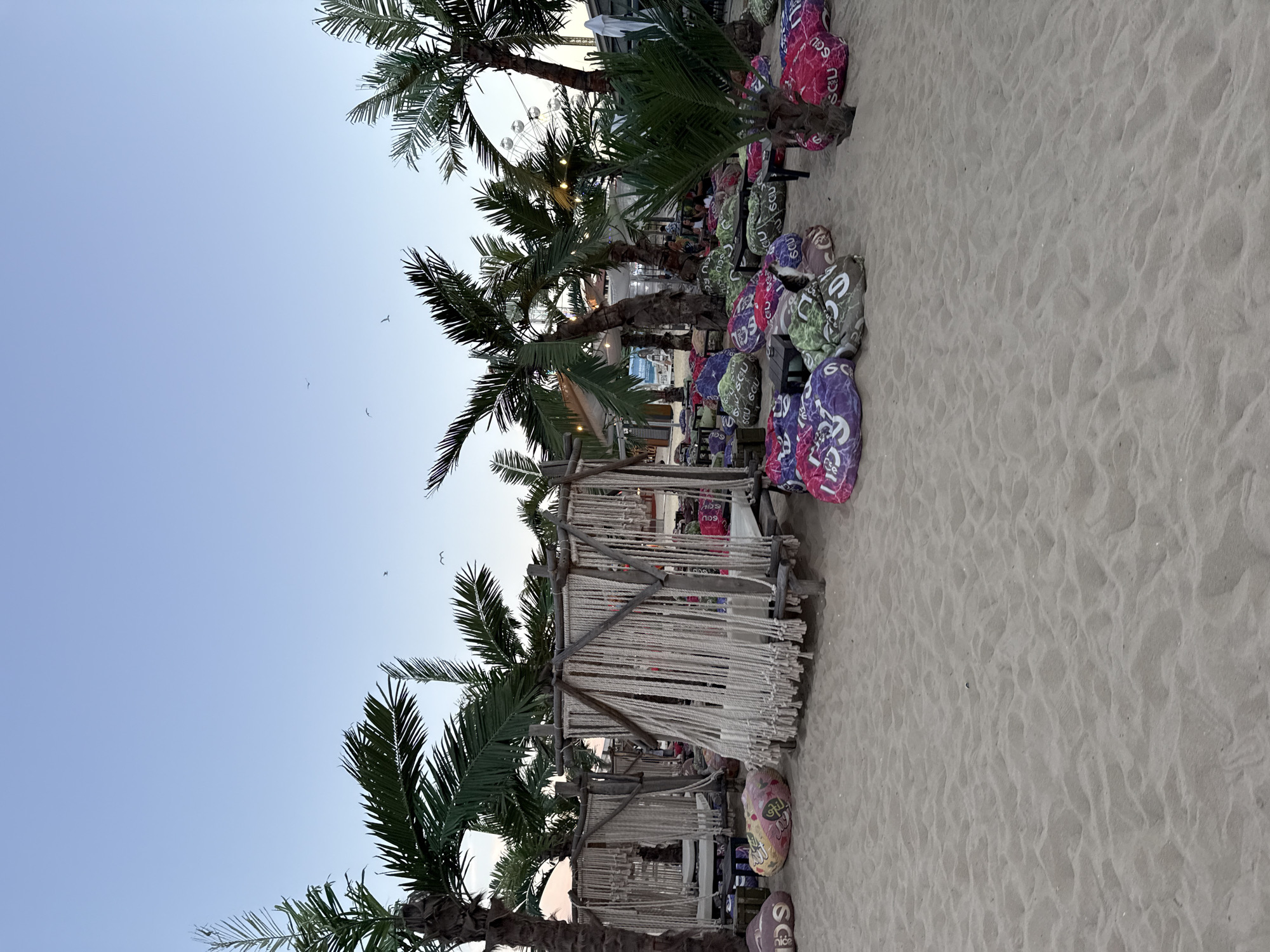 The image depicts a beach bar with palm trees and an area set up with bean bags on the sand. In the background, there is a Ferris wheel. The scene appears to be in Nesebar, Bulgaria, likely during sunset given the soft lighting and the clear sky. There are no people clearly visible in the foreground, but some individuals can be seen in the background near the bar structures. The bean bags are colorful and patterned, and some are placed around small tables. The structures in the foreground are made of wood and rope, creating a bohemian-chic ambiance. The beach is sandy, with some ripples from the tide or wind.