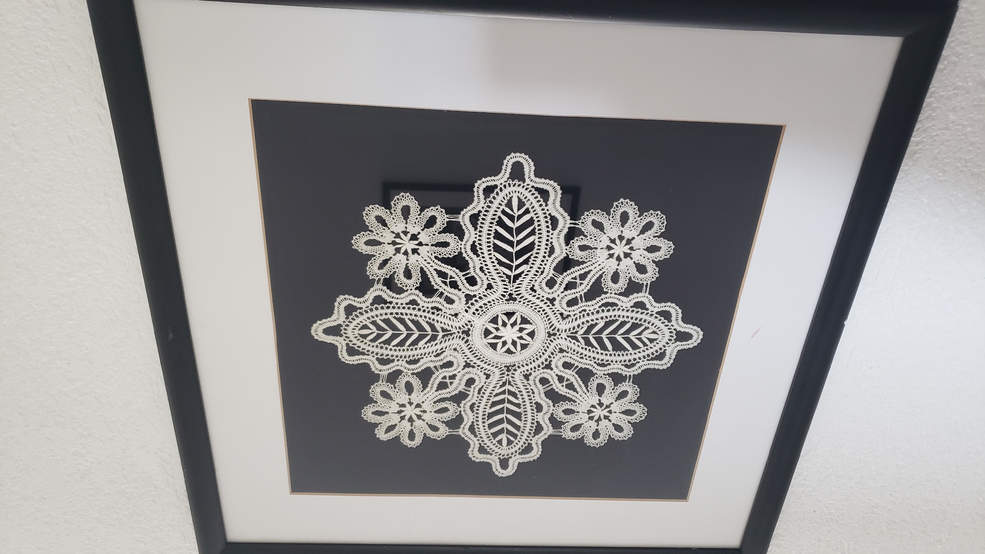 The image displays a framed piece of white lace artwork with a snowflake-like design, set against a dark gray matting. The frame is black and appears to be made of wood. The artwork is presented in a slightly angled shot, with the bottom left corner of the frame closer to the viewer. The background of the image is a textured white wall. The lace itself exhibits intricate patterns of loops, scalloped edges, and central star-like motifs. The scene is devoid of any people or explicit action. The setting is domestic, likely an interior of a home or a gallery. There is no information within the image itself to suggest a specific location in Prešov, Slovakia.