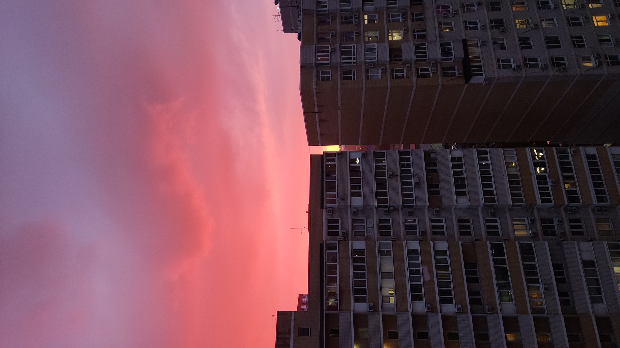 The image shows two tall buildings with a vivid red and pink sky in the background. The buildings are multi-storied and appear to be apartment blocks, based on the rows of windows and air conditioning units visible on their facades. The photograph is oriented sideways, with the buildings positioned vertically in the frame. The sky takes up a significant portion of the image, displaying vibrant colors suggestive of a sunset or sunrise. The lighting is dim, and it's possibly taken during twilight. Based on visual information and the reference, the location is suggested as Krasnodar, Russia. There are no people visible in the photograph.