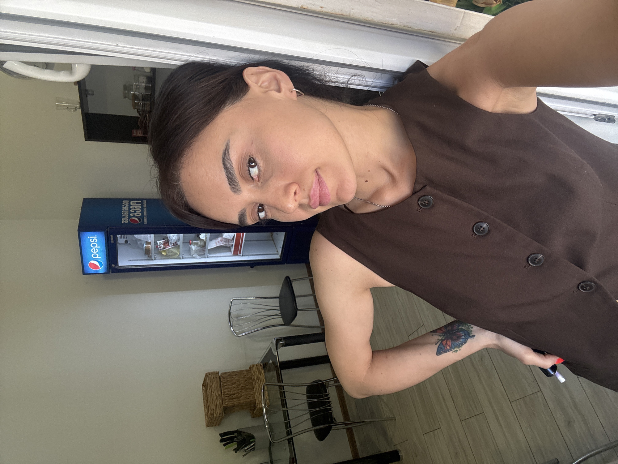 A woman with dark hair and brown eyes is taking a selfie in what appears to be a cafe or restaurant in Tbilisi, Georgia. She is wearing a brown sleeveless top with buttons down the front. She has a tattoo of a butterfly on her left forearm. In the background, a blue Pepsi refrigerator is visible, along with tables and chairs. The lighting suggests it is daytime.
