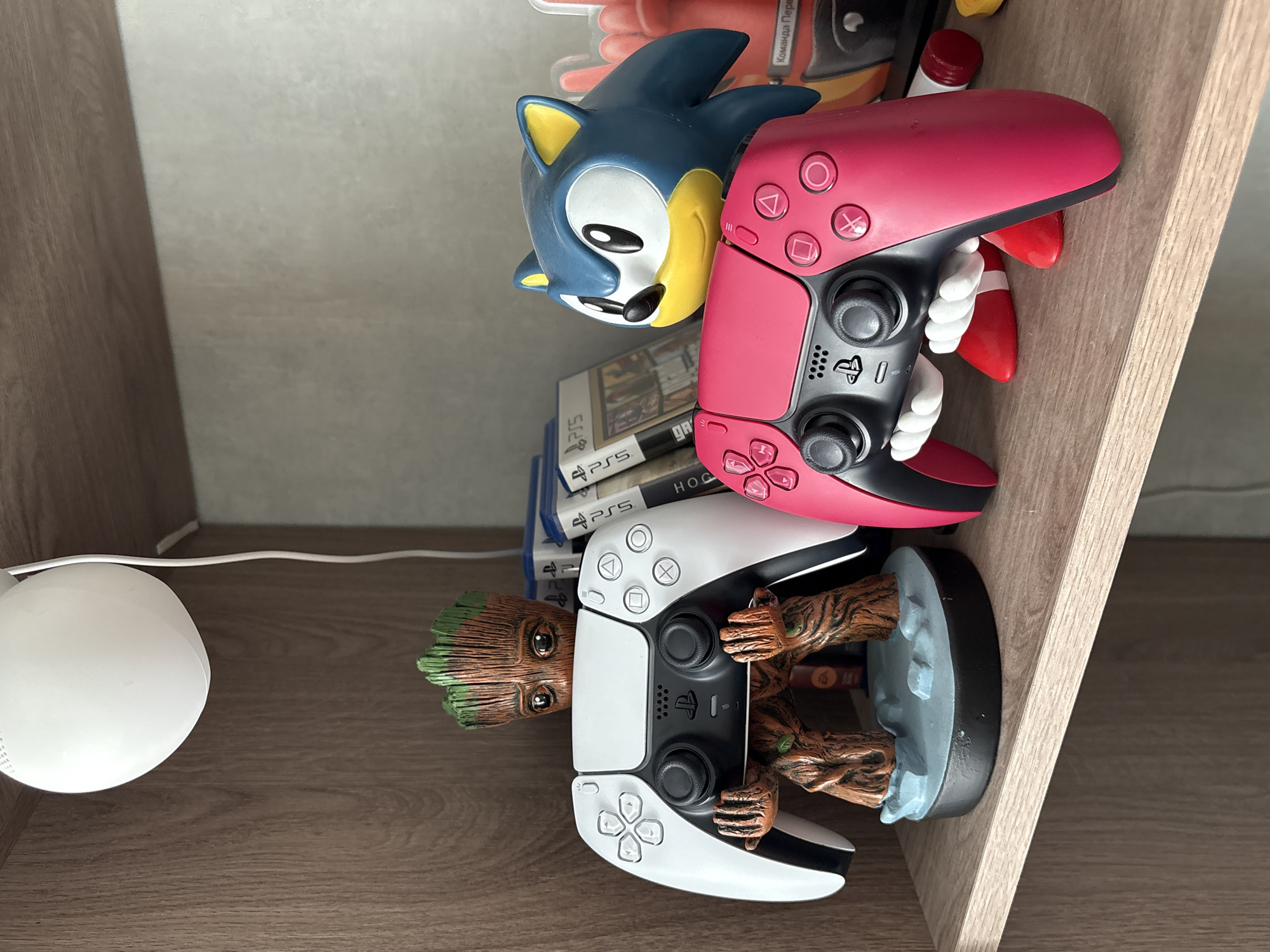 The image shows a shelf with various objects. On the bottom shelf, there is a white round lamp to the left. Next to it is a Groot figure holding a white PlayStation 5 controller. On the shelf above, there is a stack of PlayStation 5 game cases, topped by a Sonic the Hedgehog figure holding a red PlayStation 5 controller. The background is a plain wall. The shelf appears to be made of wood. The image was not taken in Birobidzhan, Russia.
