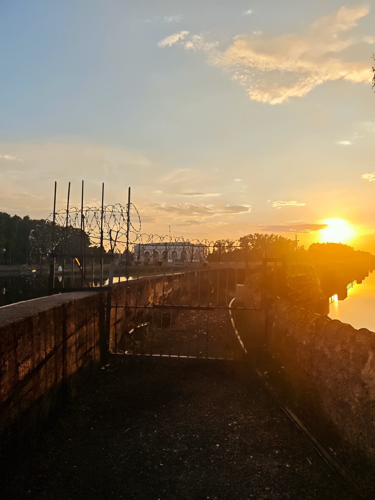 This is a medium shot of a sunset over a body of water in Vodnik SNT, Russia. The sun is a bright orb on the right side of the frame, casting a warm orange glow across the scene. The sky is a gradient of blue and orange, with scattered clouds. In the foreground, there is a weathered stone wall with barbed wire fencing along the top. A metal gate is visible in the wall. Behind the wall, trees line the water's edge. In the distance, across the water, a large building with arched windows can be seen. The water reflects the vibrant colors of the sunset. There are no people or animals visible in the image.