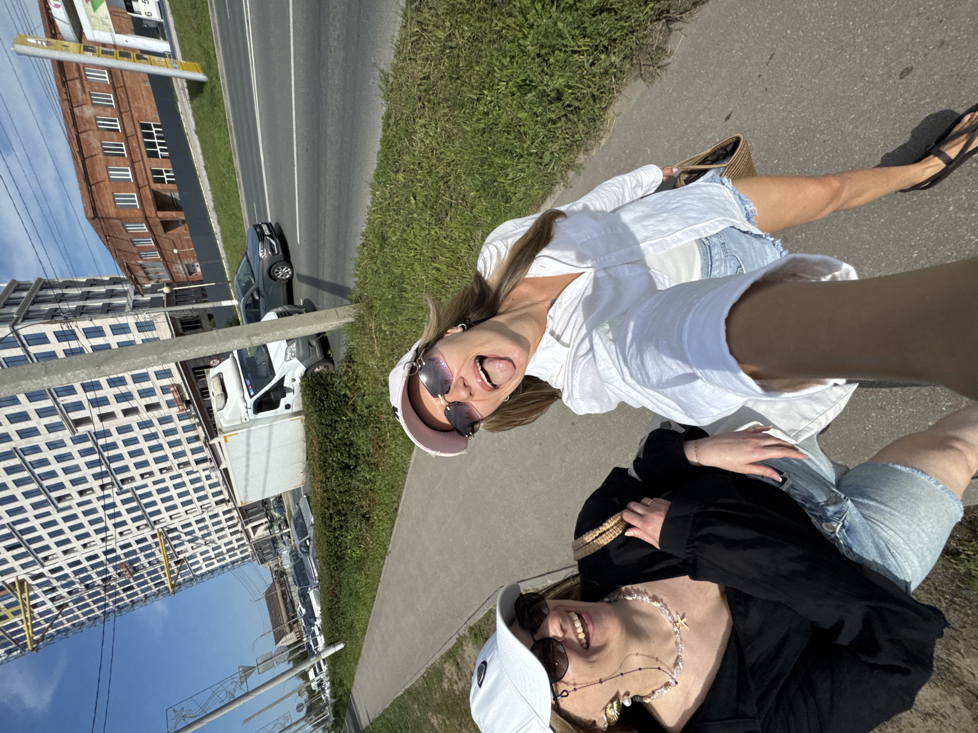 Two women are taking a selfie on a sidewalk next to a road. The woman in the foreground is wearing a black shirt, a white baseball cap, and sunglasses, and has a beaded necklace with a shell pendant. The woman behind her is wearing a white button-down shirt, denim shorts, and a pink baseball cap, and is sticking her tongue out. Both women are smiling and appear to be in good spirits.
In the background, there is a road with cars, a grassy area, and several buildings. One building appears to be an older brick structure, while another is a modern high-rise building with many windows. A white truck is parked on the side of the road. The scene is lit by bright sunlight, suggesting it is daytime. The sky is blue and clear, indicating fair weather. The overall impression is a casual outing on a sunny day.