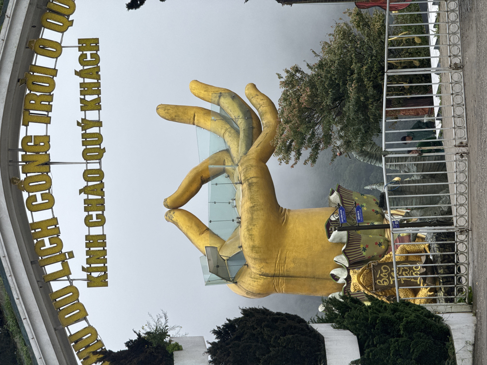 The image captures a partial view of the Golden Bridge at Sun World Fansipan Legend in Binh Lu, Vietnam. The iconic giant golden hand structure, which supports the pedestrian bridge, is prominently featured, along with the bridge itself. The surrounding environment includes trees and decorative foliage. A metal barrier or fence is visible on the right side of the frame, partially obscuring the view. Signage in Vietnamese is also seen. The sky appears overcast, suggesting foggy or cloudy weather conditions.