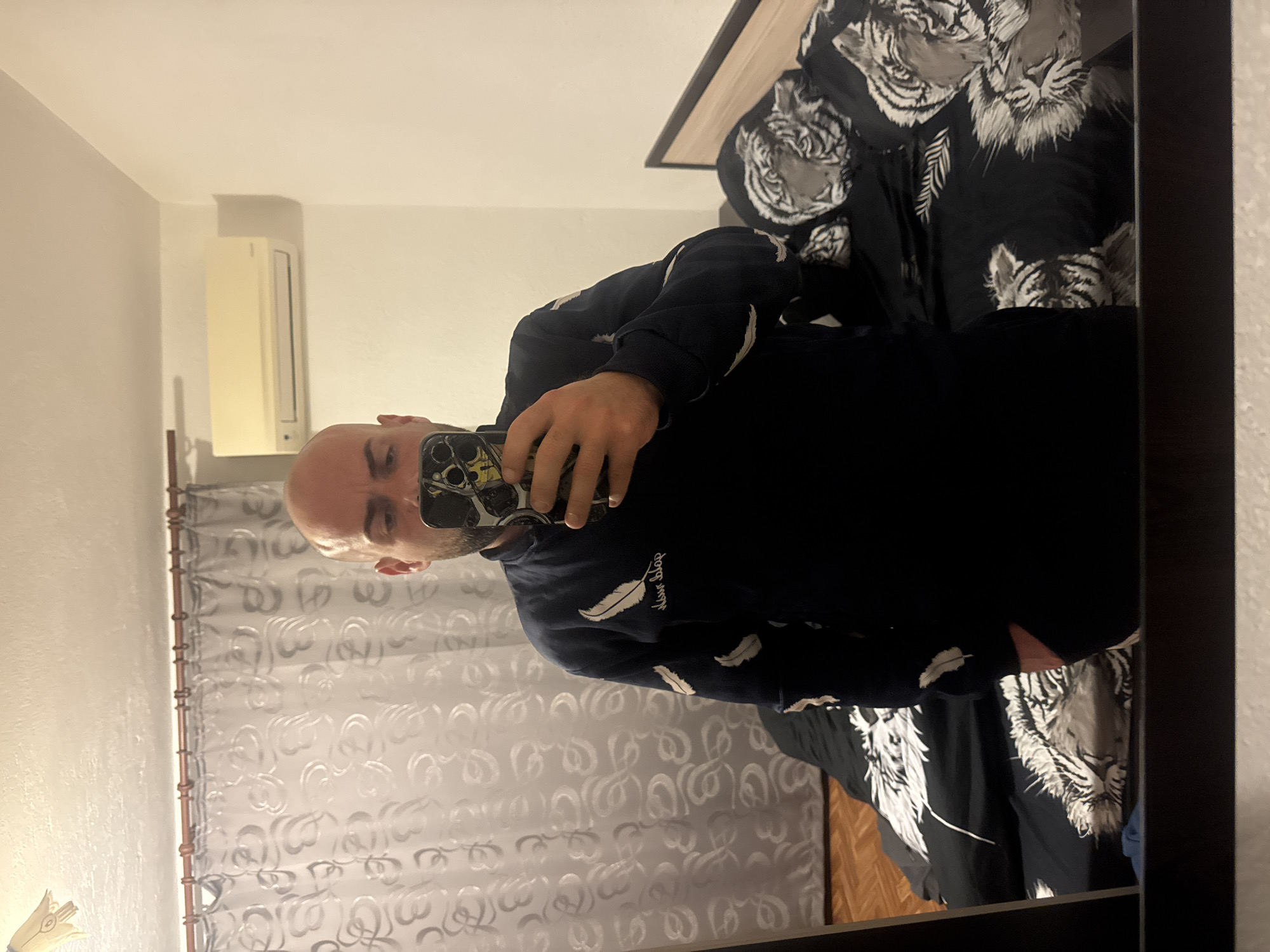 A bald man is taking a selfie in a mirror. He is wearing a black sweatshirt with white feather designs. Behind him are white curtains with a swirly pattern, and a bed with black and white tiger-print bedding. An air conditioning unit is visible on the wall. The location is Sarnevo, Bulgaria.