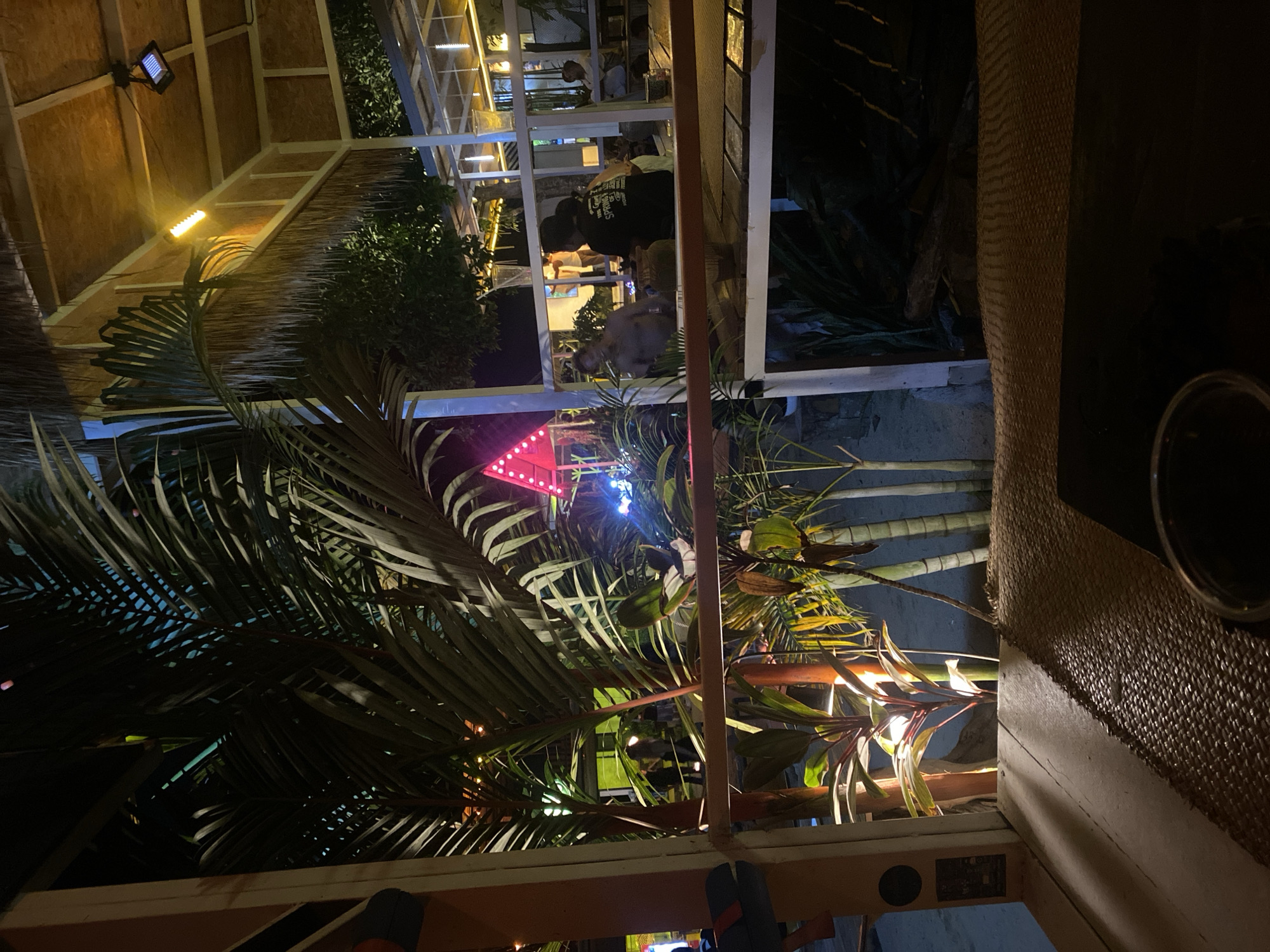 The image depicts an outdoor restaurant scene at night in Baan Hin Kong, Thailand. The perspective is from a table, with a woven placemat visible. A palm tree is prominent in the foreground, its leaves partially obscuring a view of the restaurant's open-air structure. String lights and a red triangular sign are visible. Patrons are present in the restaurant area.