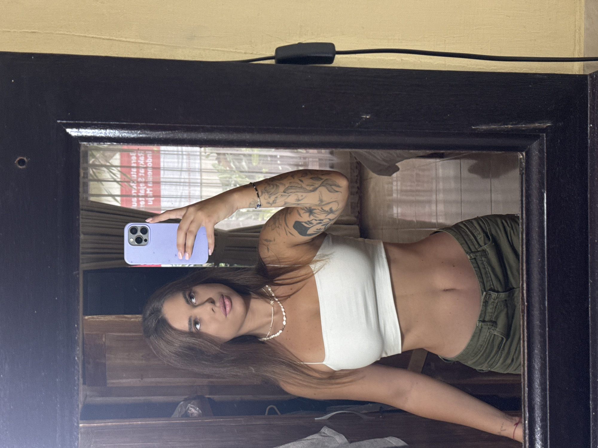A young woman with brown hair is taking a mirror selfie. She is wearing a white crop top, olive green pants, and two necklaces. She has tattoos on her left arm. She is holding a light purple phone with a case. The mirror is framed in dark wood and is reflecting a room with wooden doors and a window. The wall behind the mirror is yellow.