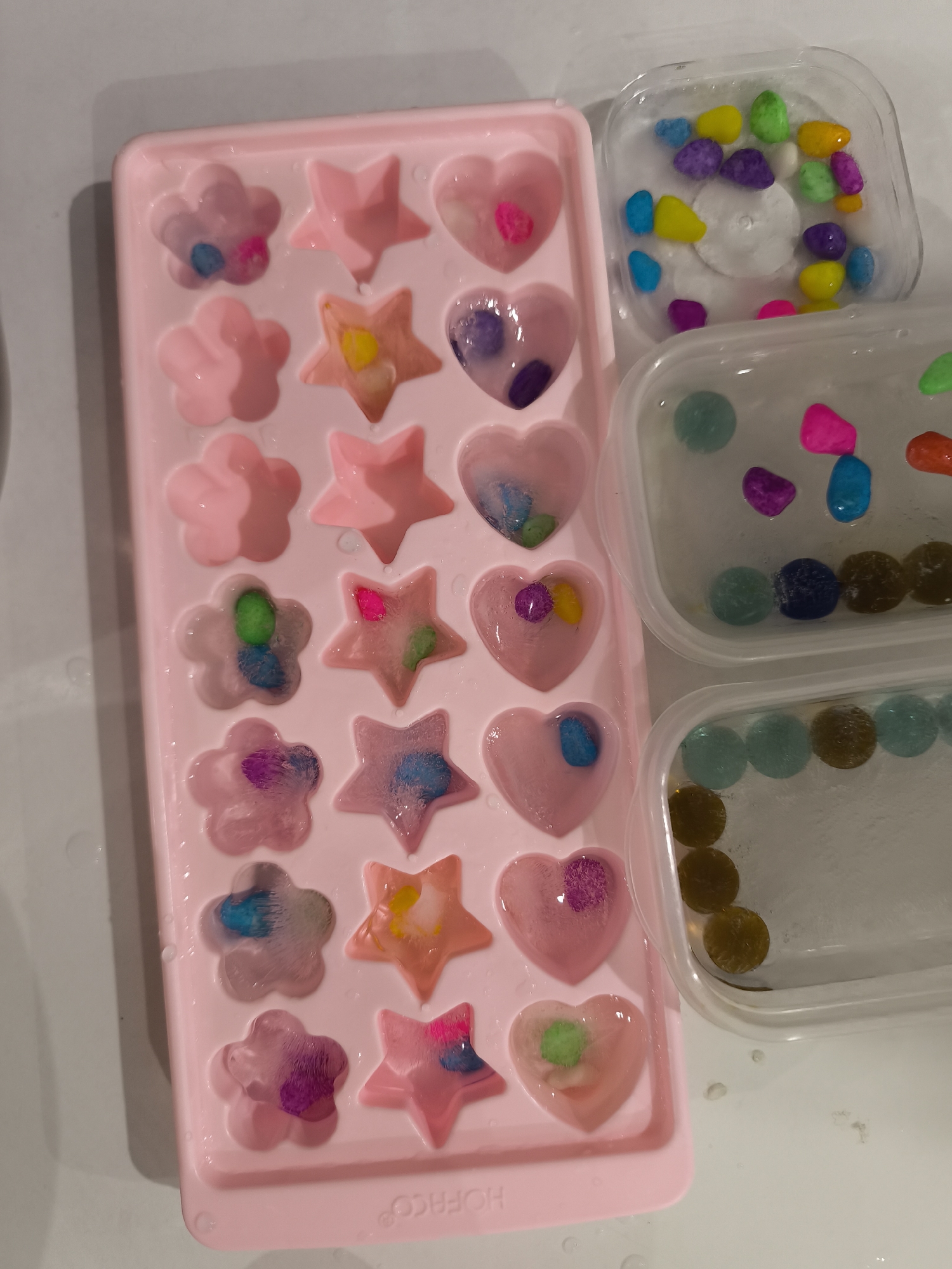 The image displays a pink silicone ice cube tray filled with ice in the shapes of flowers and stars, with colorful candy pieces embedded within some of the ice cubes. To the right of the tray are two clear plastic containers also filled with ice and candy pieces. The background appears to be a white surface, possibly a countertop. There are no people or animals visible in the image. The scene is indoors. The provided context about the location in the city of Bac Nha Trang Ward, Vietnam, cannot be verified from the image itself.