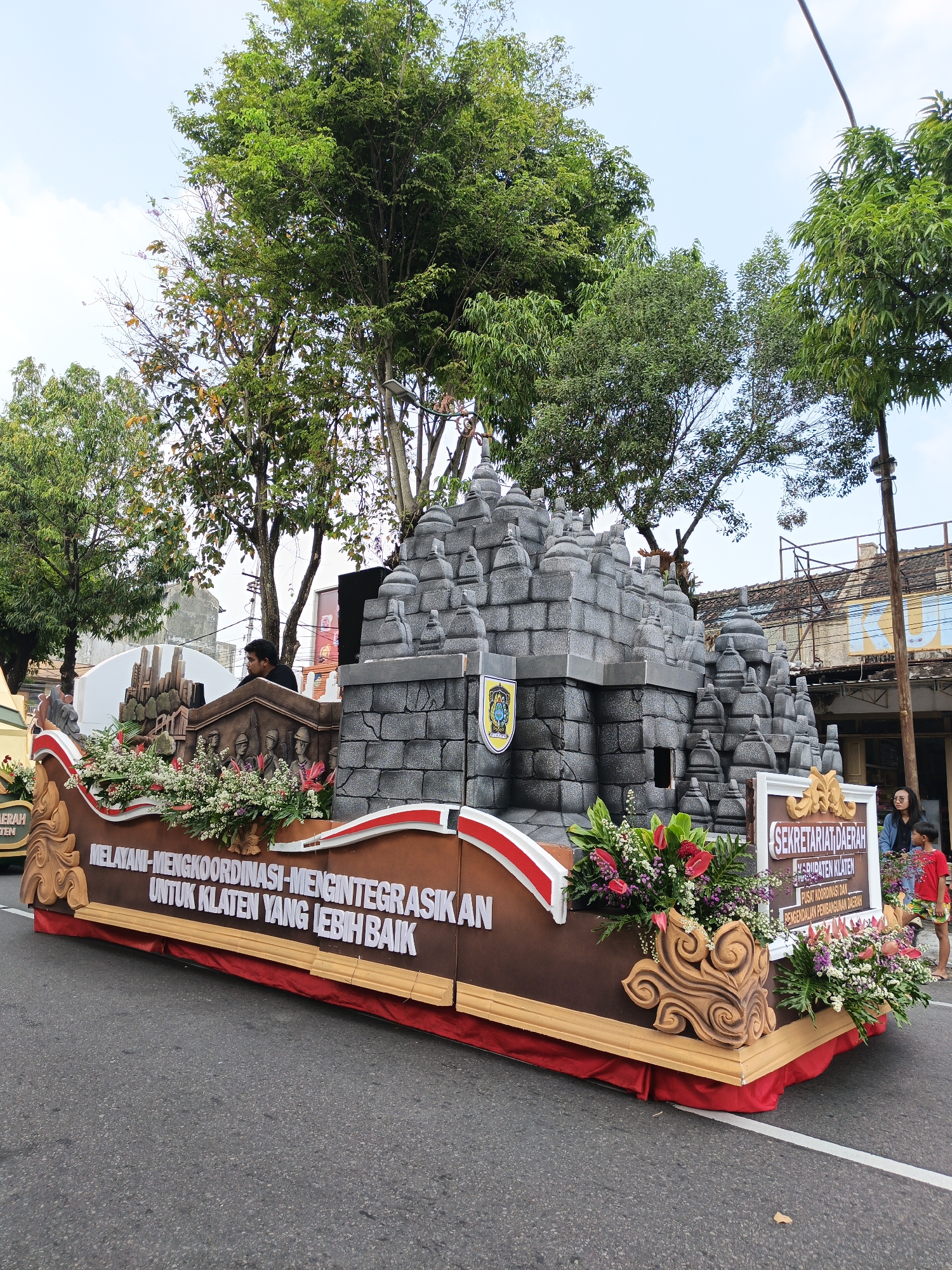 The image shows a parade float on a paved road in Klaten, Indonesia. The float is decorated with a replica of a stone temple structure, surrounded by flowers and foliage. A large banner on the side of the float reads, "MELAYANI-MENGKOORDINASI-MENGINTEGRASIKAN UNTUK KLATEN YANG LEBIH BAIK" (Serving-Coordinating-Integrating for a Better Klaten). A sign on the float also indicates it represents the "SEKRETARIAT DAERAH KABUPATEN KLATEN" (Regional Secretariat of Klaten Regency). Several people are visible, including a man standing behind the float, a woman and a child on the right side, and other individuals in the background. Trees line the street, and buildings are visible further down the road. The sky is clear and blue.