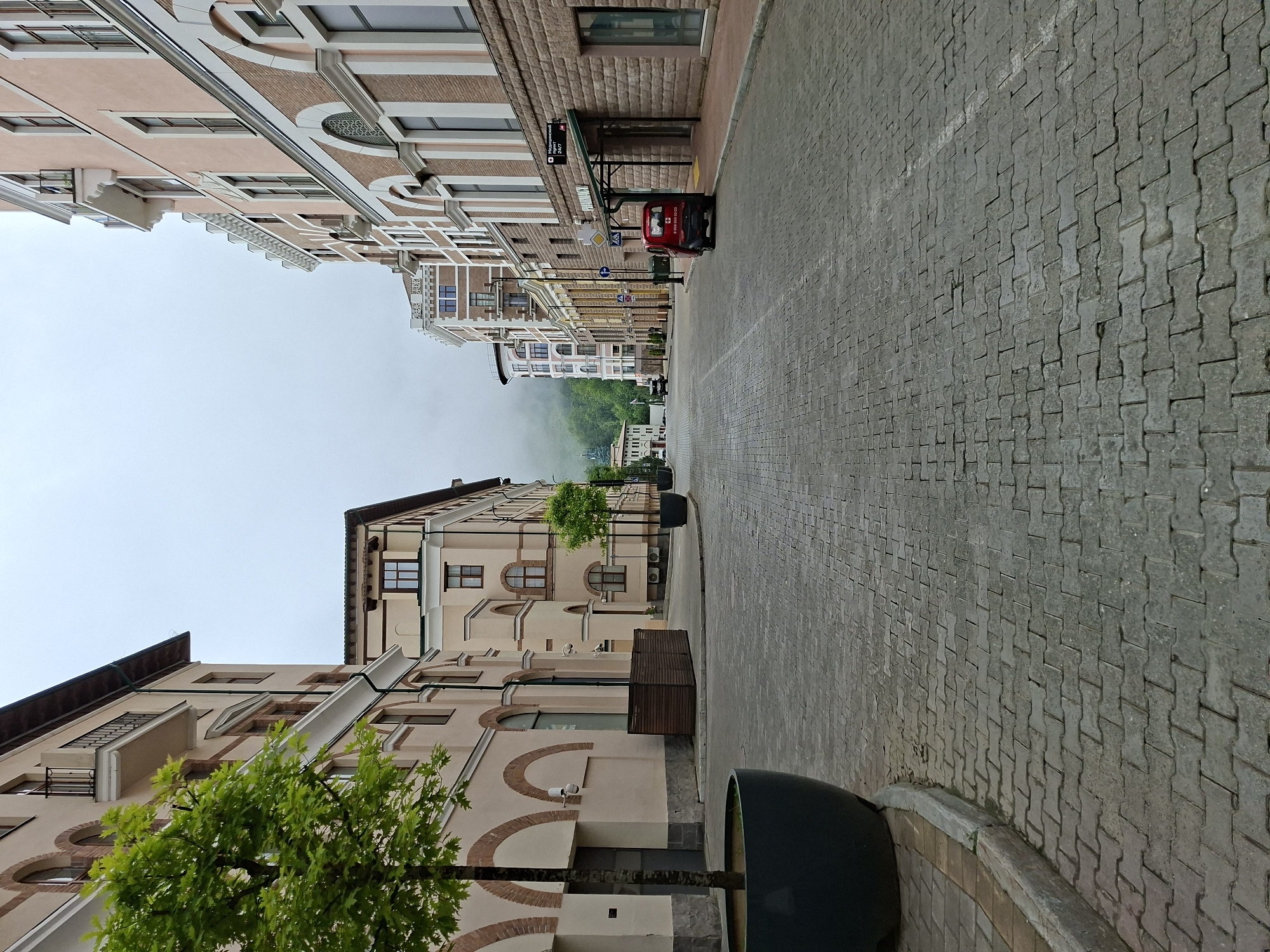 The image depicts a street scene in Estosadok, Russia, characterized by cobblestone paving and multi-story buildings with light-colored facades and decorative brickwork. A large tree in a dark planter is visible on the left, with smaller trees lining the street on the right. A red vehicle is parked on the right side of the street, near a building with signage indicating a "PHARMACY". The sky is overcast, and a misty mountainous landscape is visible in the background. The overall atmosphere is calm and quiet, suggesting an early morning or a less busy time of day.