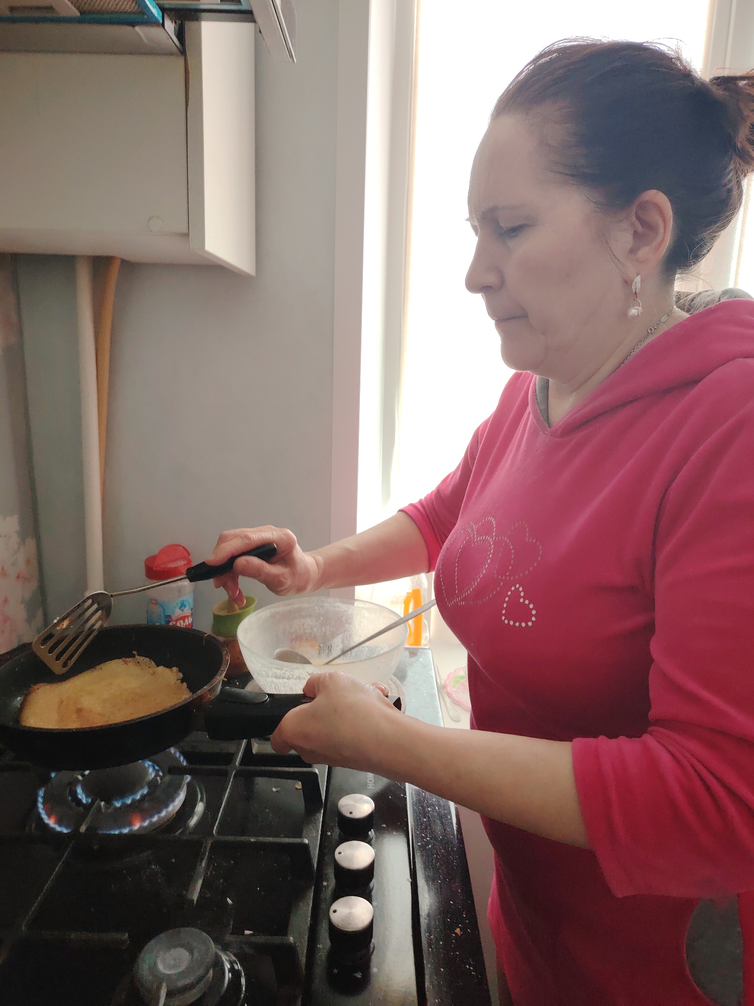 In an indoor kitchen setting, a middle-aged woman with dark hair pulled back into a bun stands facing left. She wears a pink hooded sweatshirt adorned with heart designs. The woman is actively engaged in cooking. She holds a black frying pan over a lit gas burner on a black stovetop. Inside the pan is a golden-colored pancake or crepe. She uses a metal spatula in her left hand to manipulate the food. Beside the stovetop is a clear glass bowl and other kitchen items, including a spice container, a spoon and an orange canister. A white kitchen cabinet is visible on the left, along with pipes. A brightly lit window with vertical blinds is positioned behind her. The location is likely a home kitchen, and given that no specific clues regarding the location in Neftekamsk, Russia are identifiable, this information is unavailable.