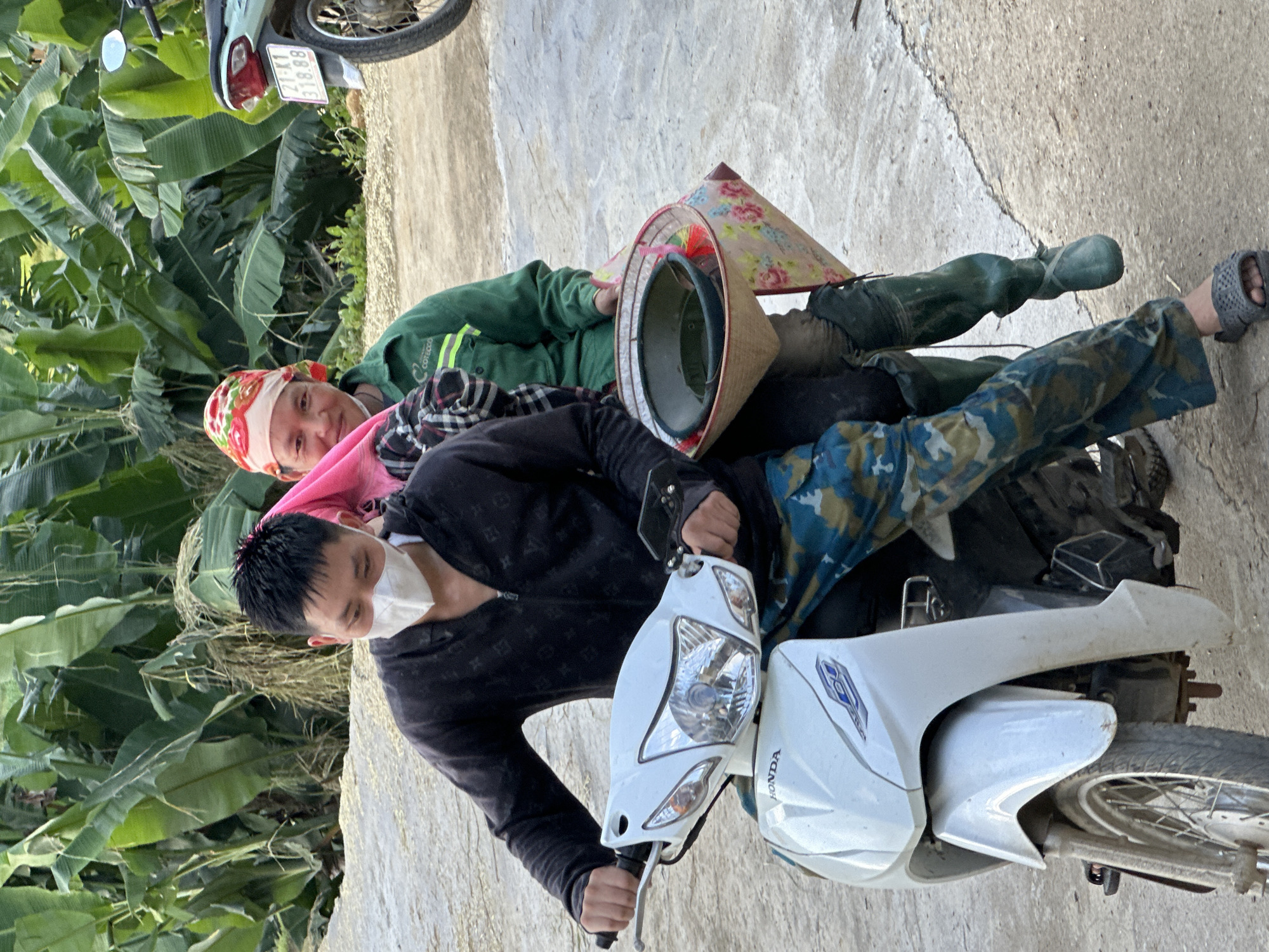 In Gia Hội, Vietnam, an outdoor scene depicts two individuals on a white Honda scooter on a concrete path. A male driver, wearing a black zip-up hoodie with faint patterns, camouflage pants, dark sandals, and a white medical face mask, operates the vehicle. His hands are on the handlebars. Behind him, a female passenger with a red and white floral bandana, a green long-sleeved shirt featuring a 'COTECCO' logo, and dark green waders or boots, sits with a slight smile, looking towards the camera. She carries a traditional Vietnamese conical hat (nón lá) lined with pink floral fabric, which contains a dark green bowl or basin. The background consists of dense green foliage, consistent with banana plants. A light-colored scooter with a license plate '21-K1 318.88' is partially visible on the right side of the frame.