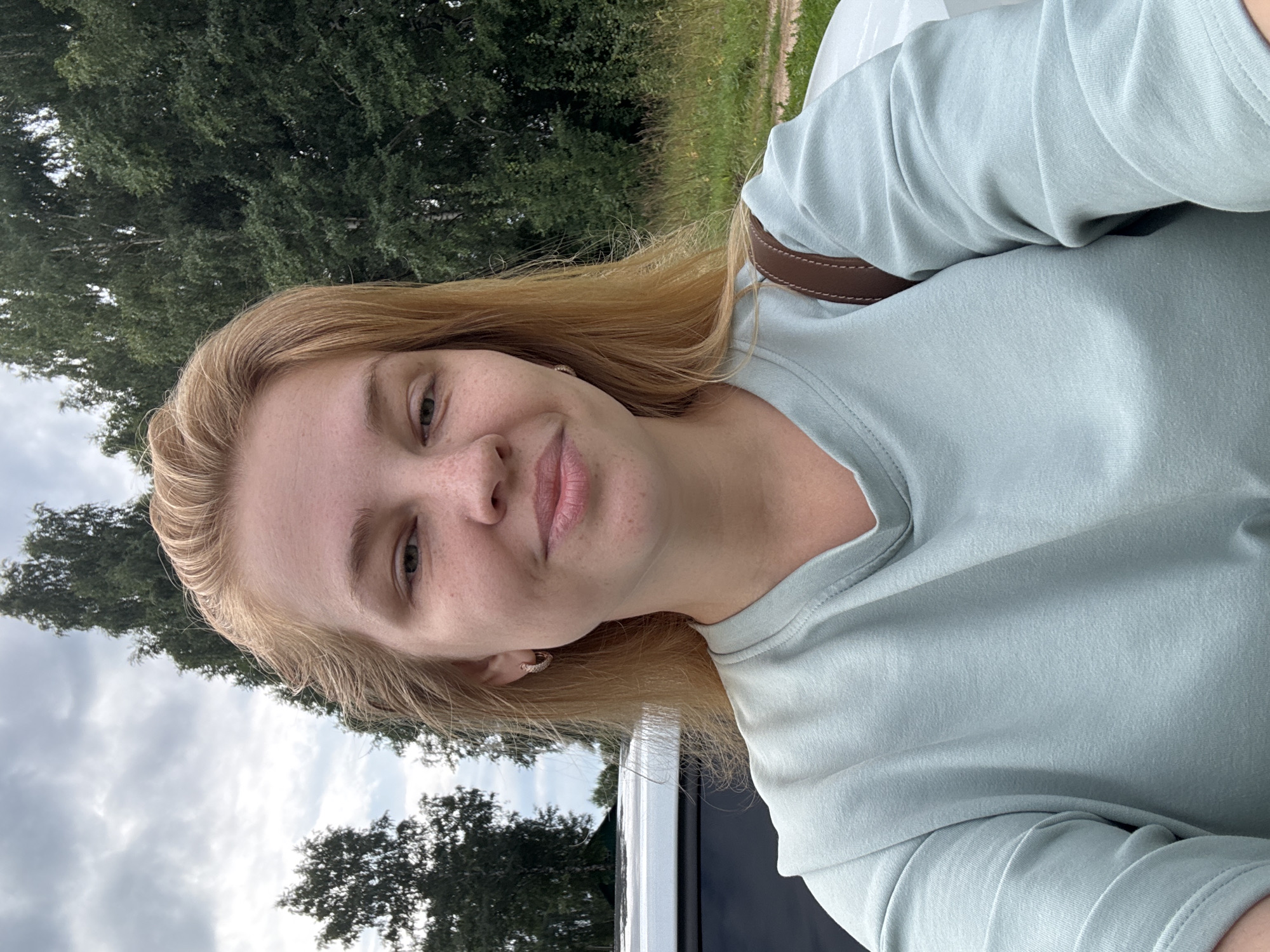 A young Caucasian woman with shoulder-length blonde hair and freckles takes a selfie outdoors. She is wearing a light blue crewneck t-shirt and a brown strap crosses her shoulder. Behind her, a white car is partially visible, along with a backdrop of lush green trees and a cloudy sky. The location appears to be Cherkasovo, Russia.