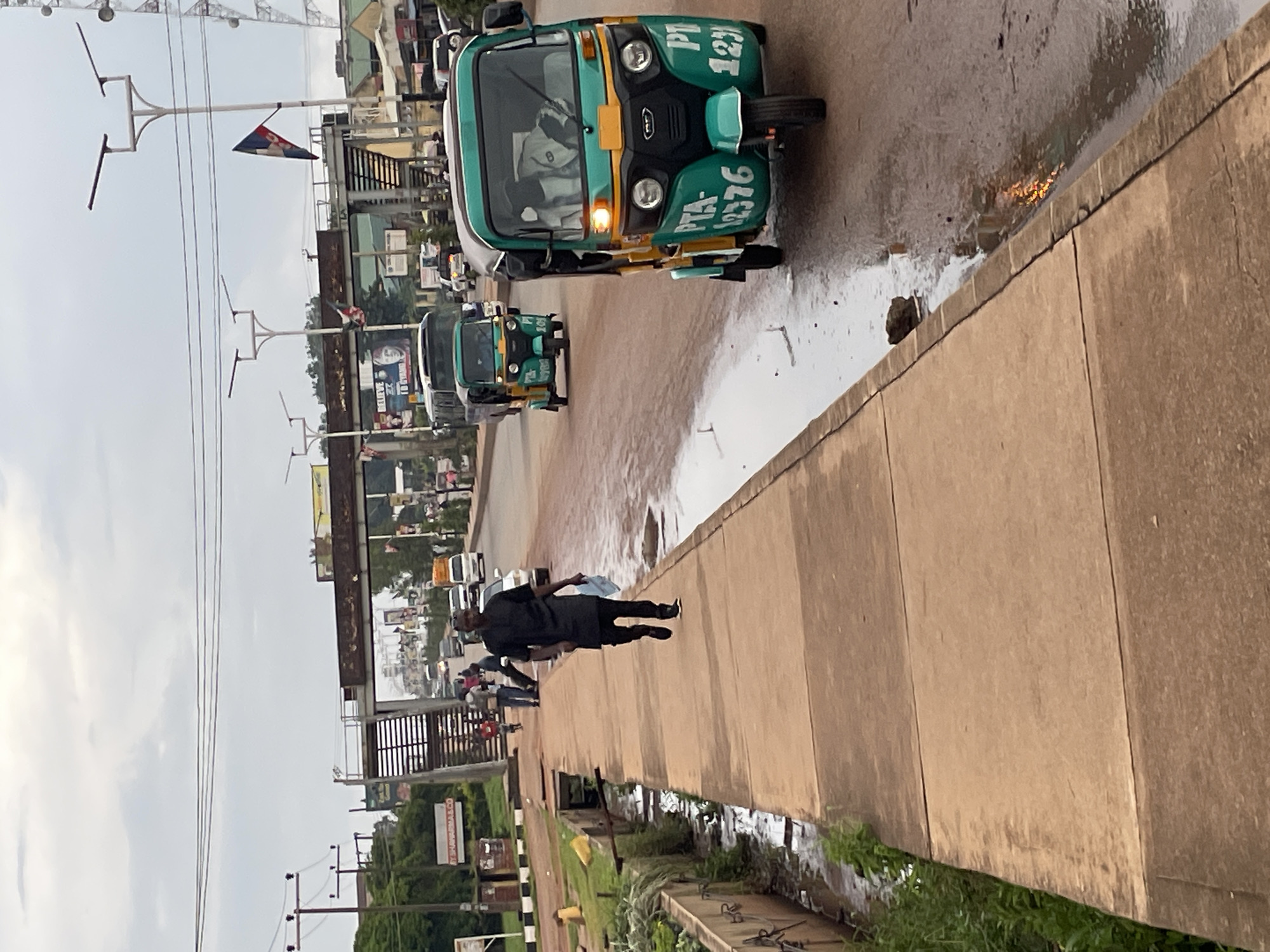The image depicts a street scene in Jos, Nigeria, with a prominent focus on a pedestrian walkway and the adjacent road. A man dressed in dark attire is walking on the concrete walkway, carrying a light-colored item in his left hand. Two auto-rickshaws, known locally as Keke Napep, are visible on the road. The front auto-rickshaw is green and yellow and displays the license plate "PTA-12376". The second auto-rickshaw, also green, is positioned behind it. Several other vehicles, including a bus and cars, are present in the background, suggesting moderate traffic. Pedestrian bridges and lampposts are also visible, along with some overhead wires. The weather appears overcast. The environment shows signs of wetness on the road surface, indicating recent rain. Vegetation can be seen along the edge of the walkway and in a drainage channel. Billboards and signs are present on buildings in the distance.