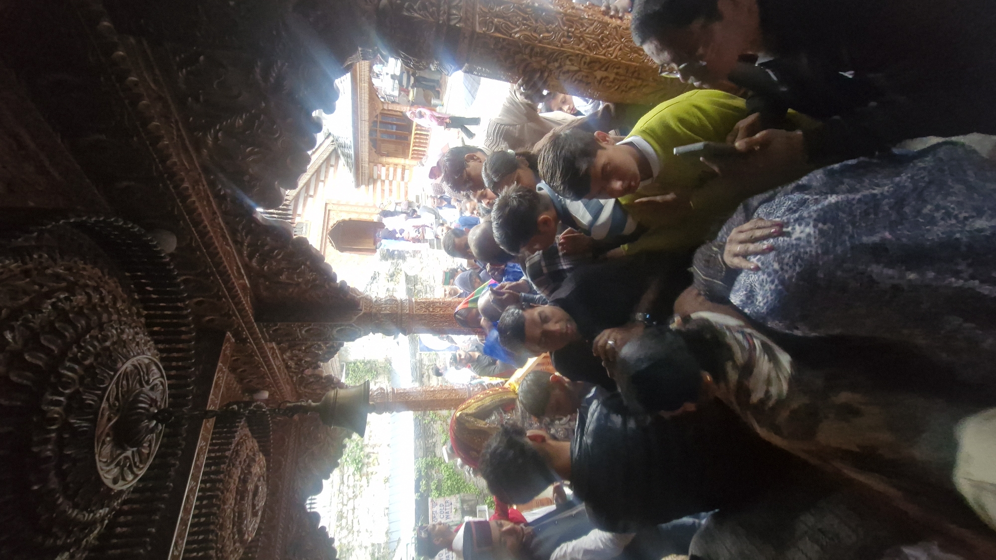 The image captures a densely crowded scene inside a wooden structure with intricate carvings. In the foreground, a group of people are gathered, many looking towards the center where a decorated effigy or shrine is visible, covered in colorful fabric. A bell hangs from the wooden ceiling. The crowd appears to be engaged in a religious or celebratory event. In the background, more people are present, some standing near buildings and pathways. The overall atmosphere suggests a public gathering in a cultural or spiritual setting within Vashisht, India.